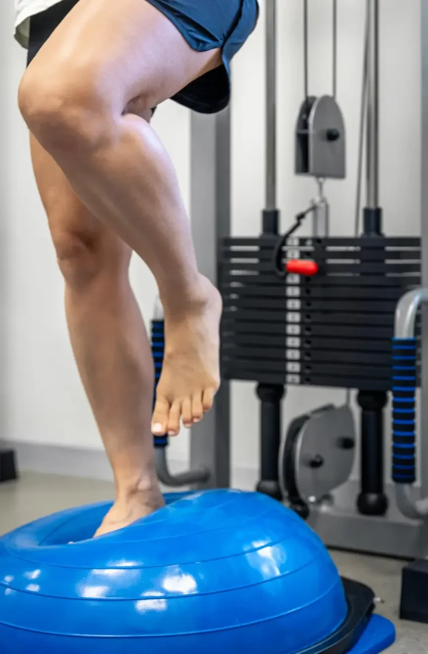 Person balancing on a blue wobble cushion in a gym, with gym equipment in the background.