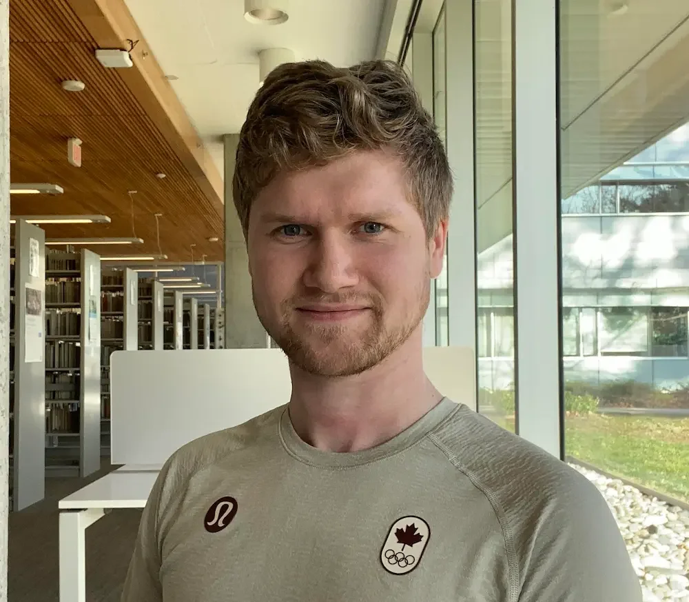 A man with light brown, wavy hair and a light beard standing indoors near large glass windows in a modern building, wearing a gray athletic shirt with a Lululemon logo and a Canadian Olympic Team badge.