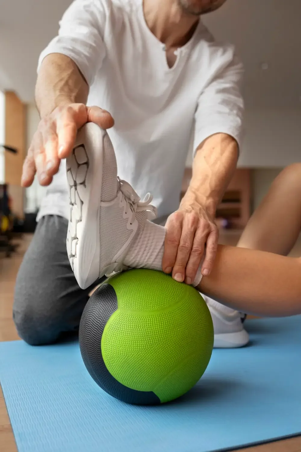 Physical therapist assisting with balance exercise using a green and black massage ball, with a person wearing white athletic shoes and white socks on a blue exercise mat.