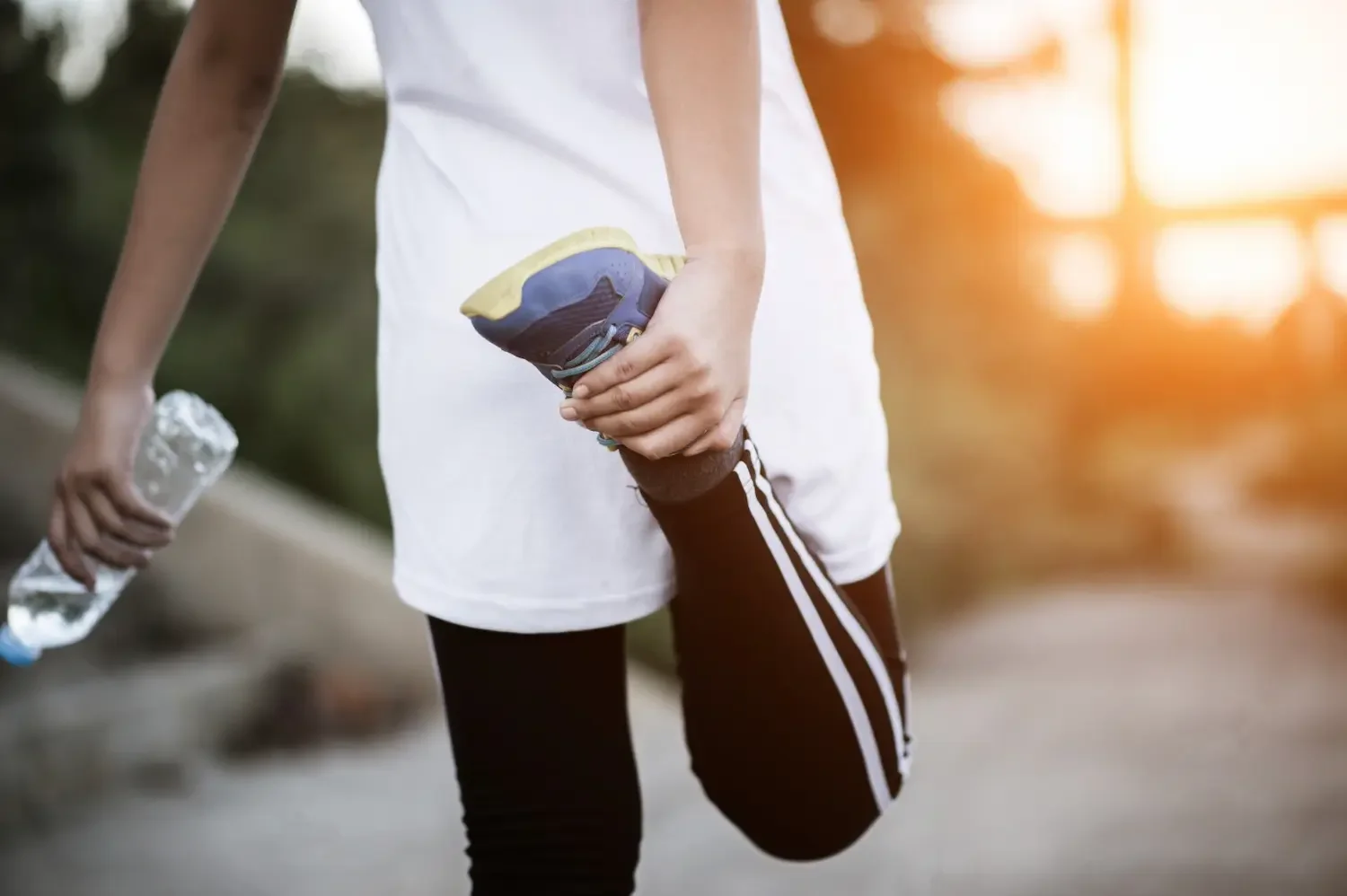 Person in athletic clothing stretching their quadriceps muscle while holding a water bottle and a shoe in an outdoor setting at sunset.