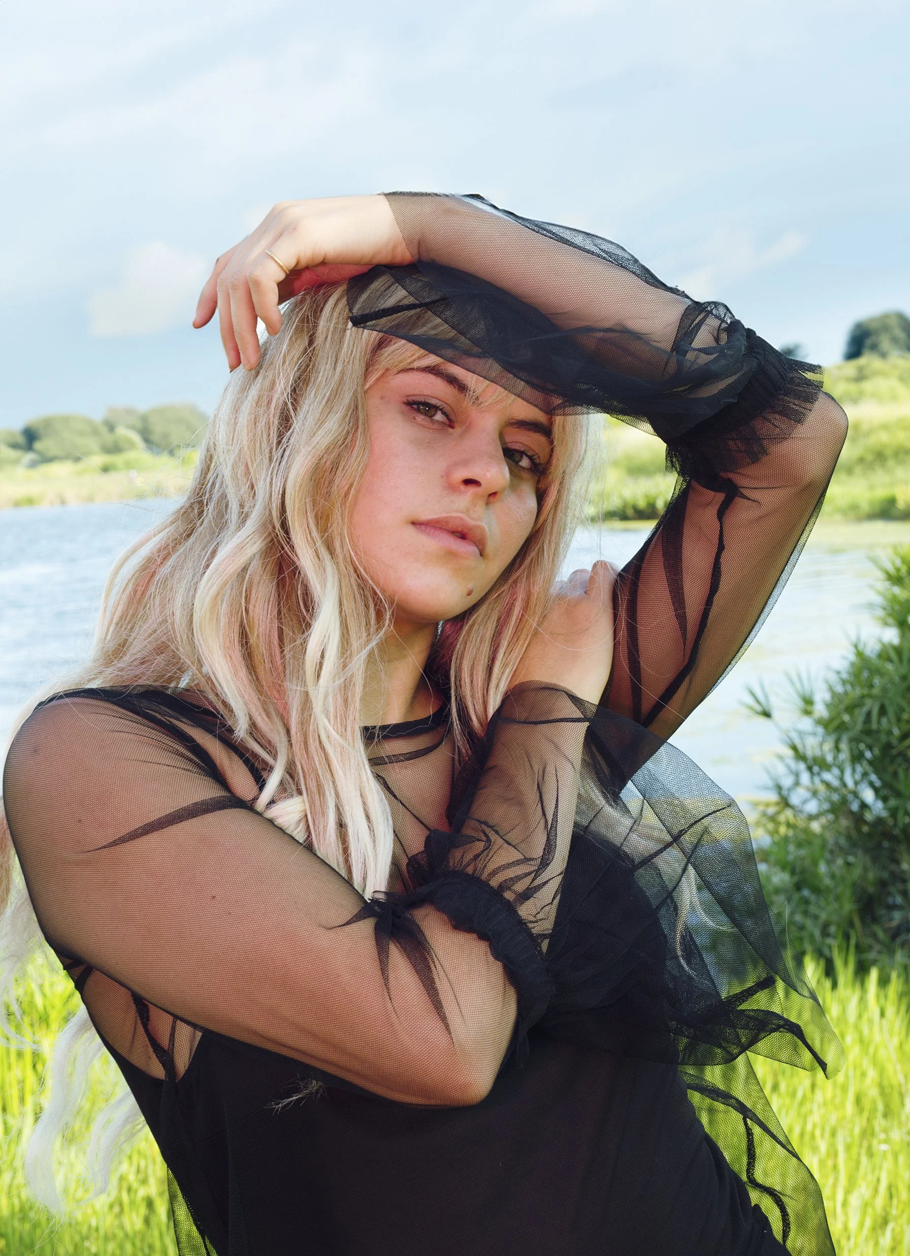 Young woman with blonde hair wearing a black sheer dress, standing outdoors near a body of water with green grass and trees in the background, under a blue sky with some clouds.