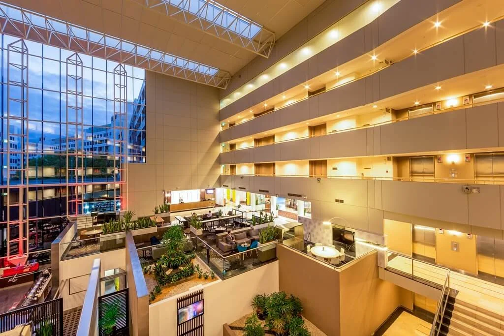 Interior view of a modern hotel lobby with multiple floors, large glass windows, greenery, and seating areas.