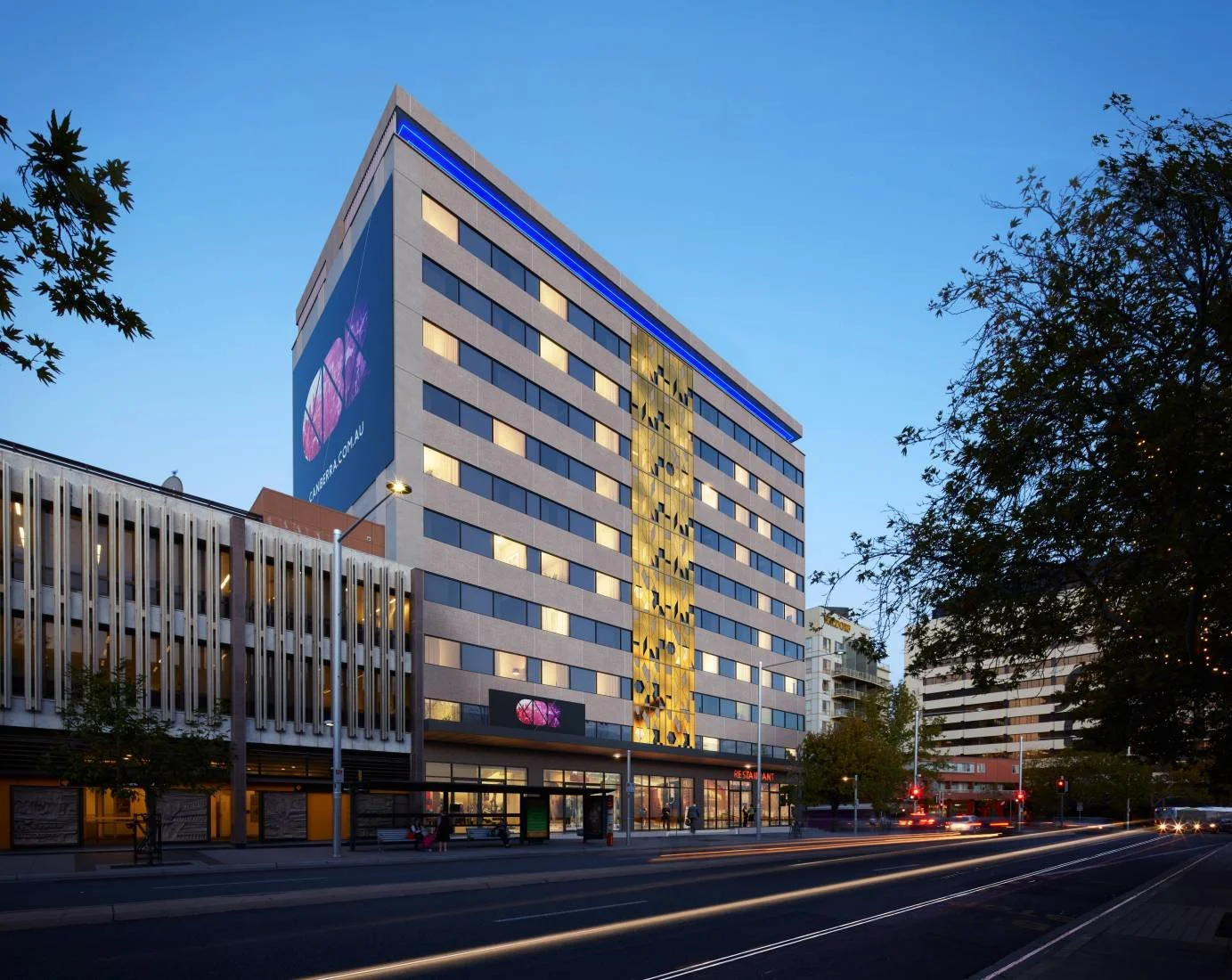 A modern multi-story building with a purple and yellow geometric design on its facade, illuminated signs, and a street with light trails from moving vehicles at dusk.