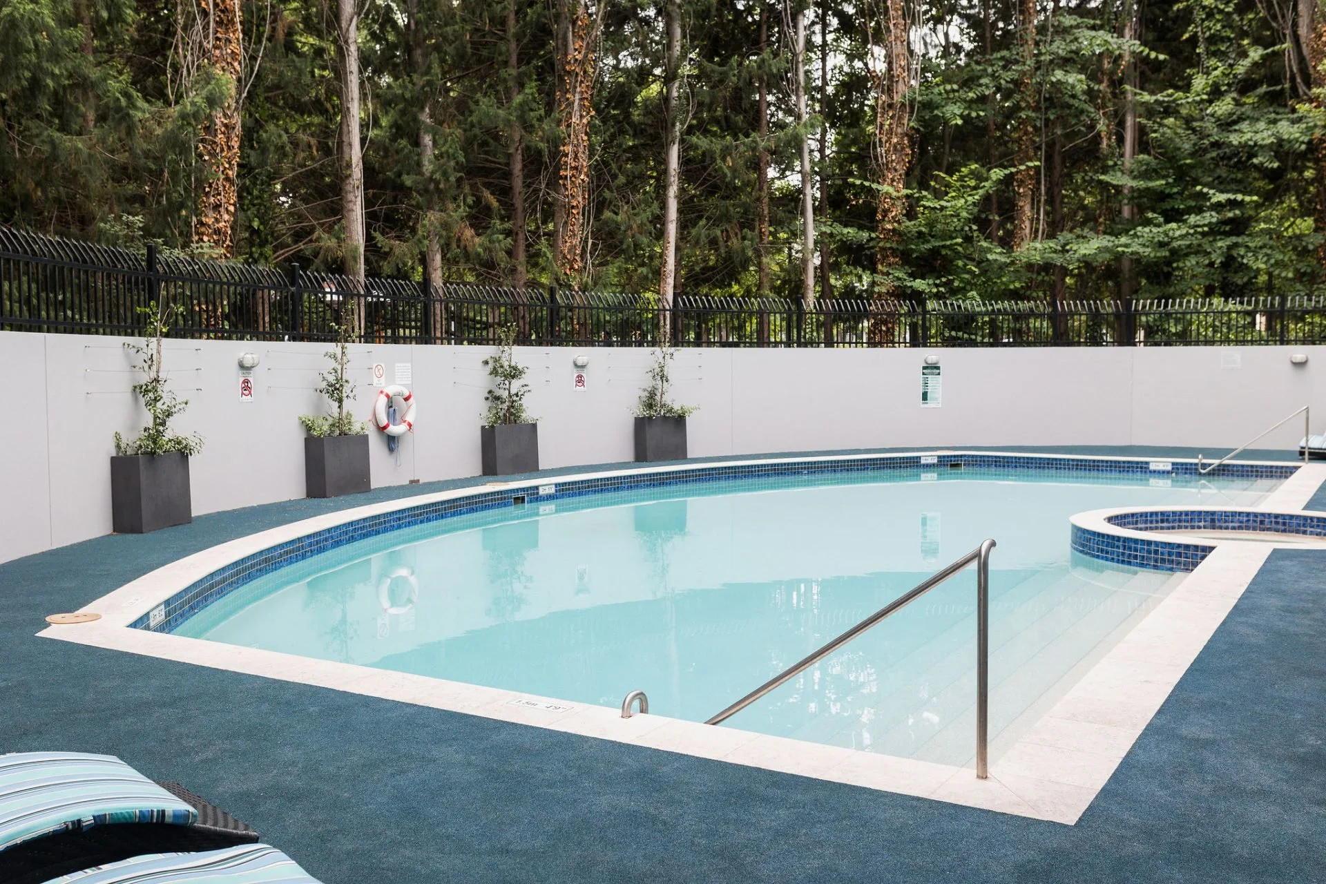 Empty rectangular swimming pool surrounded by a blue deck, with a forest of trees in the background. Pool has metal handrails and a small circular hot tub attached on the right side. There are potted plants and safety equipment along the white wall around the pool.