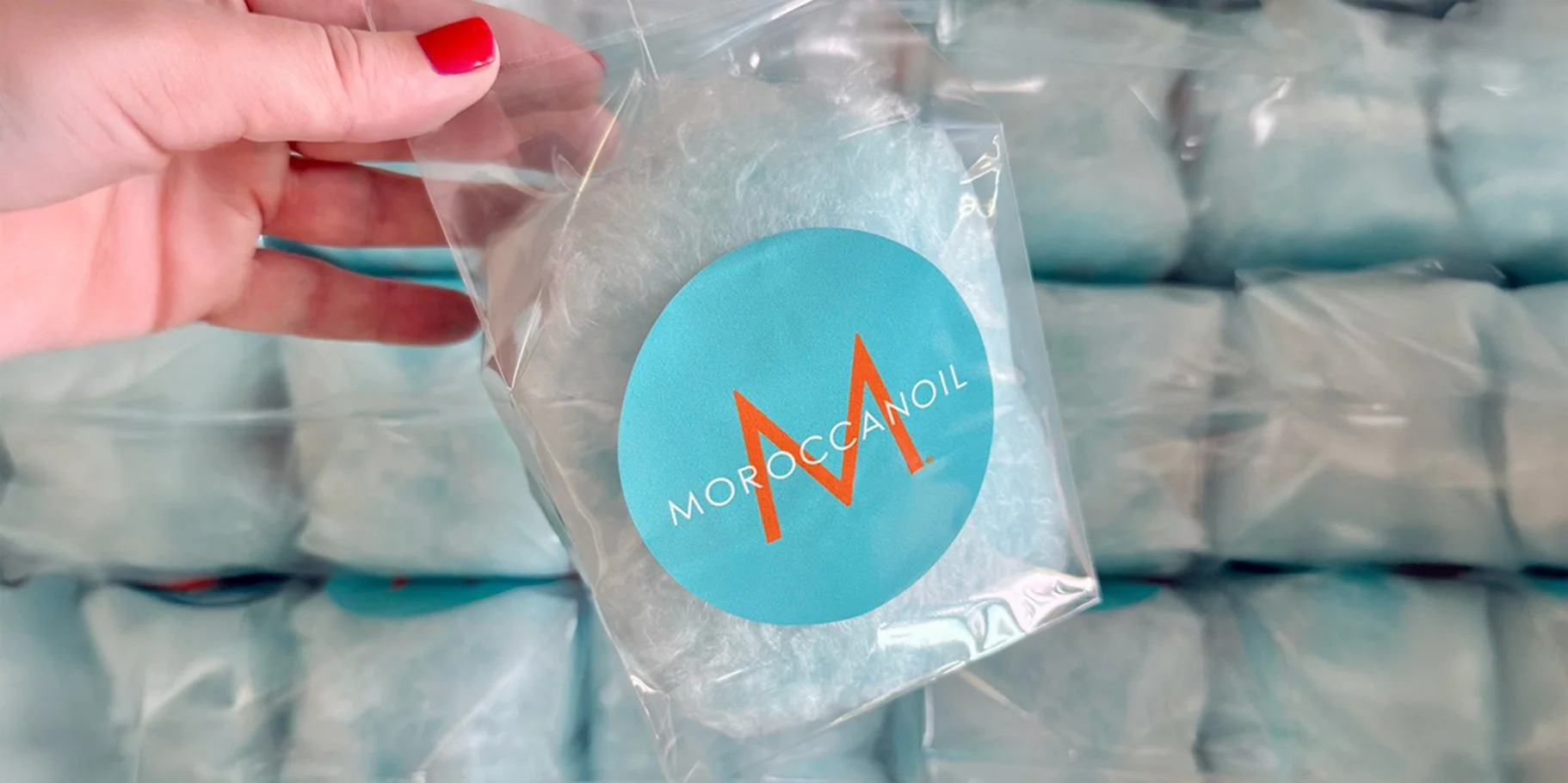A hand with red nail polish holding a clear plastic bag with a blue circular label that has an orange logo and white text reading 'Moroccan Oil' against a background of stacked blue packages.