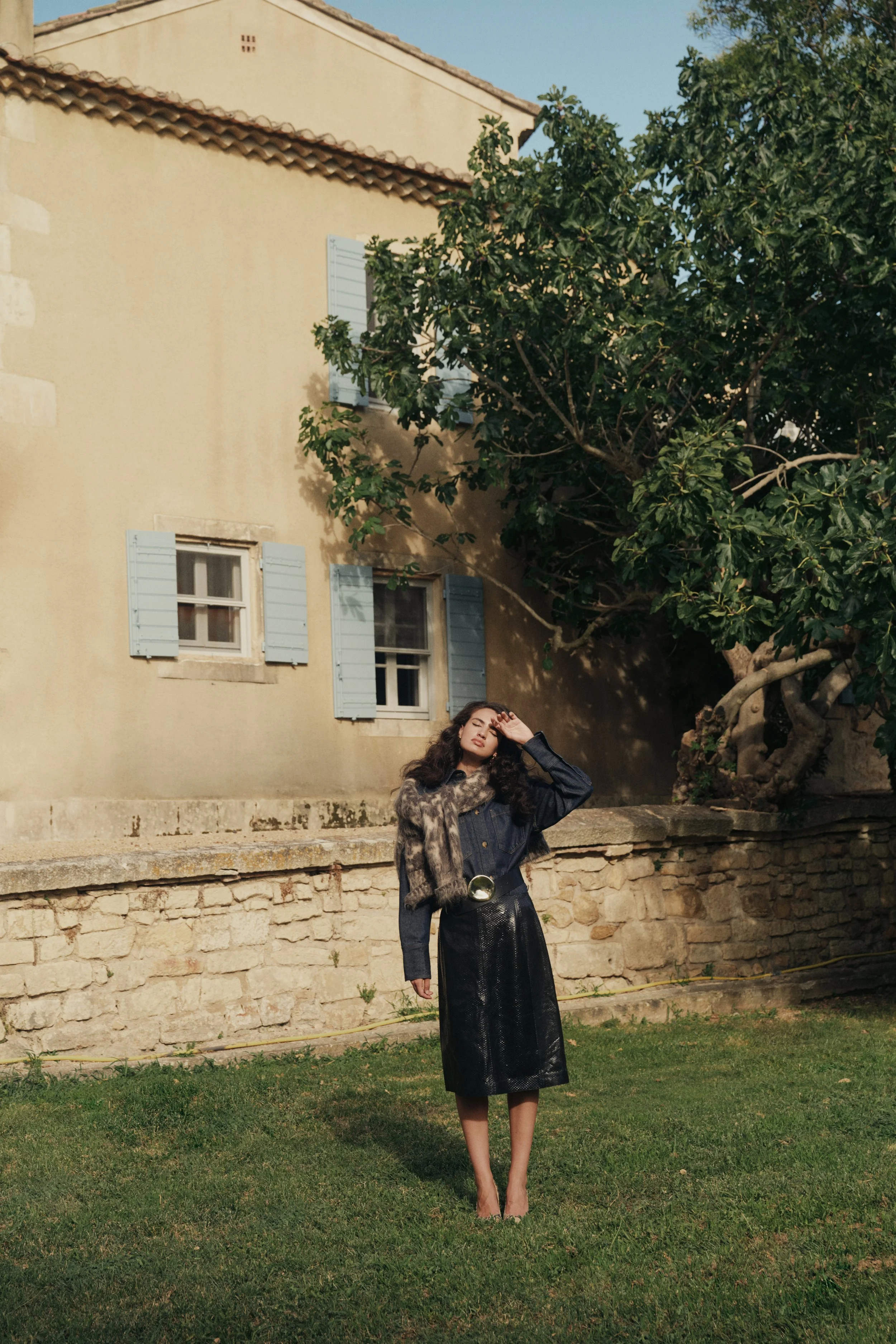 A young woman with curly hair standing on a grassy area in front of an old stone wall, a beige house with blue shutters, and a large tree, during daytime. She is wearing a black leather skirt, a denim jacket with fur, and is shielding her eyes from the sun with her hand.