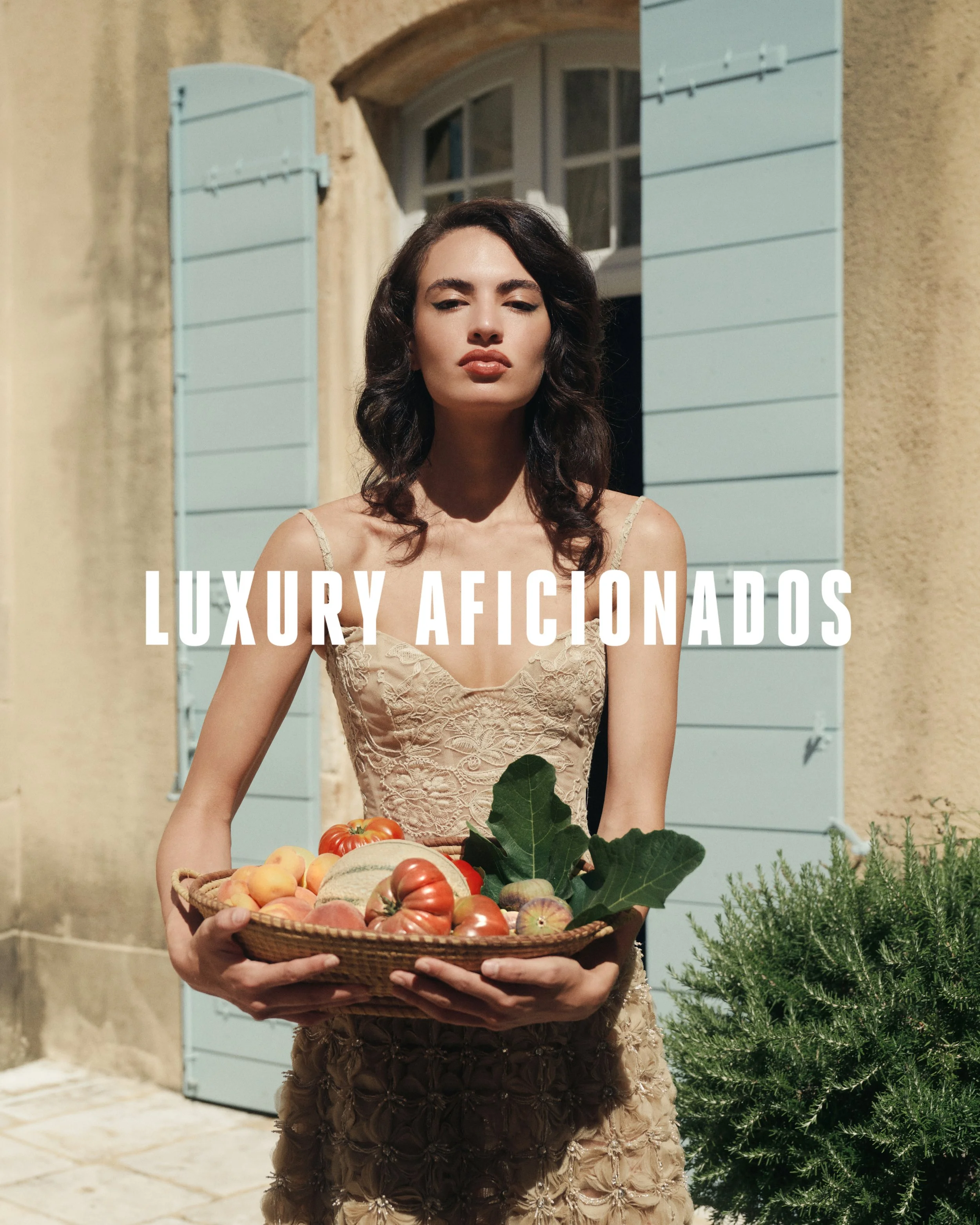 A woman in an elegant, beige, floral-patterned dress holding a basket of assorted fresh vegetables and fruits, standing outdoors in front of a building with light blue shutters and a window, with a green bush on the side.
