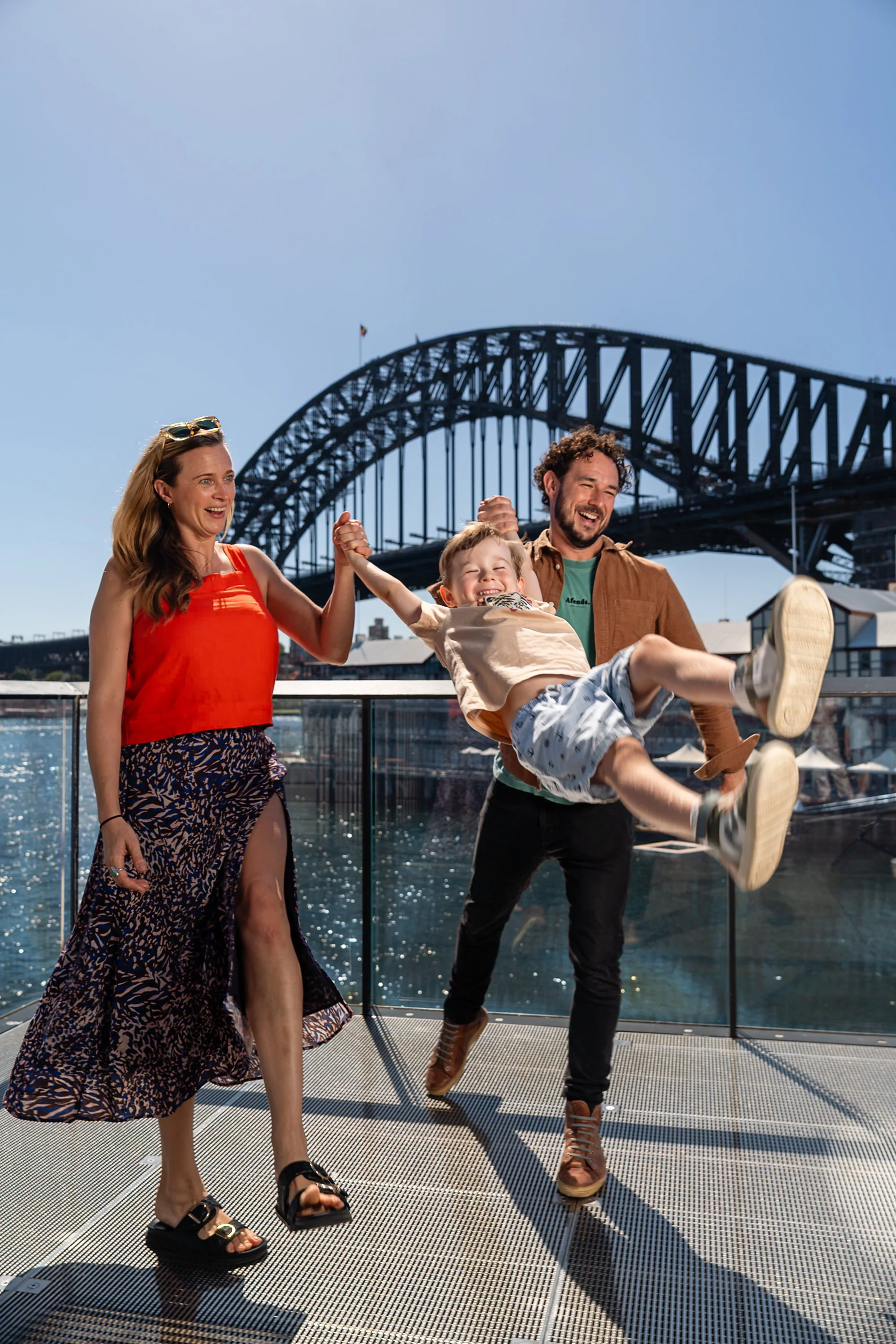 A family of three celebrating on a boat deck with the Sydney Harbour Bridge in the background. The woman is wearing a patterned skirt and red top, the man is wearing a brown jacket and green shirt, and their young child is in a beige shirt and shorts