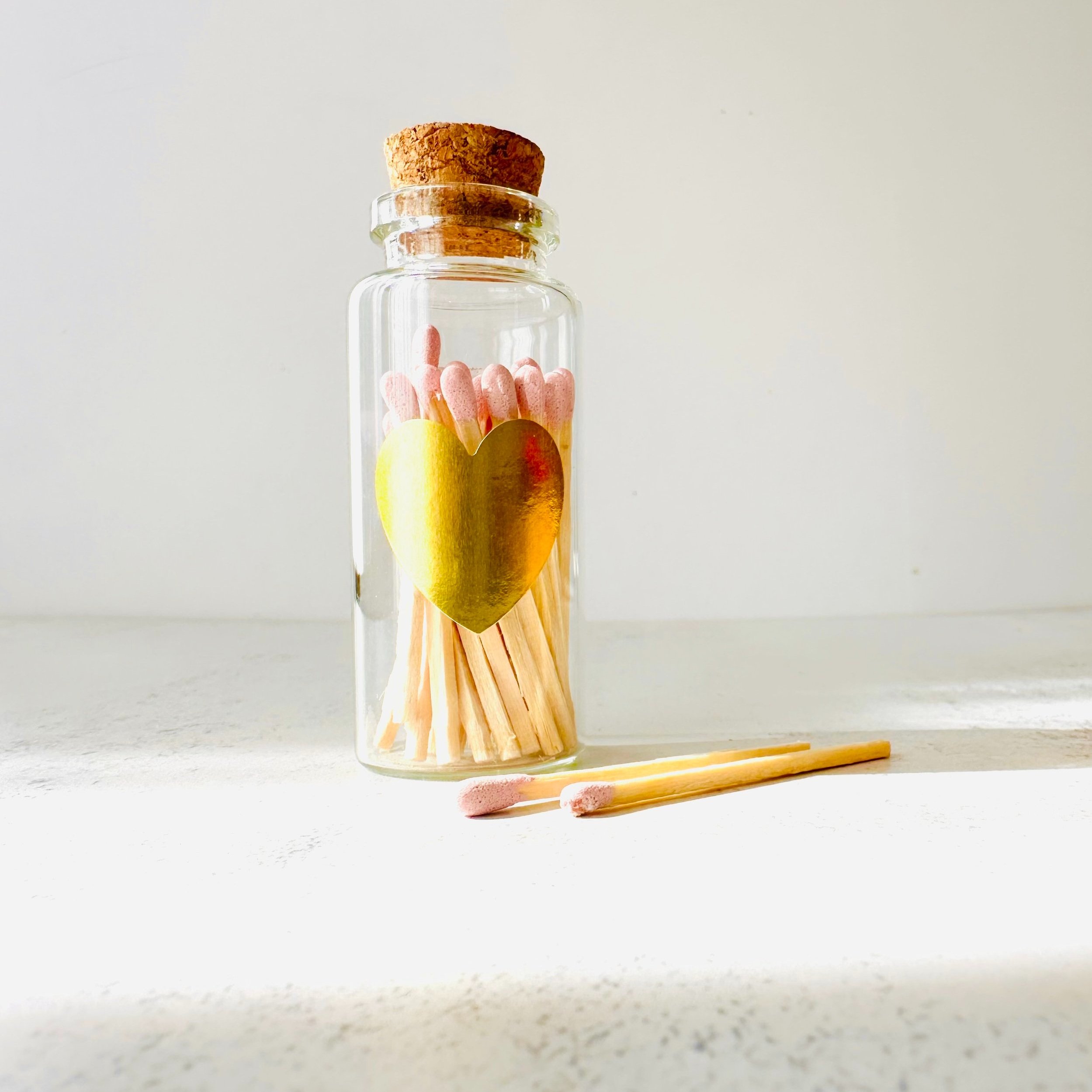 Glass jar with pink-tipped matches and a cork lid, featuring a gold heart sticker, two matches on the side, white background.