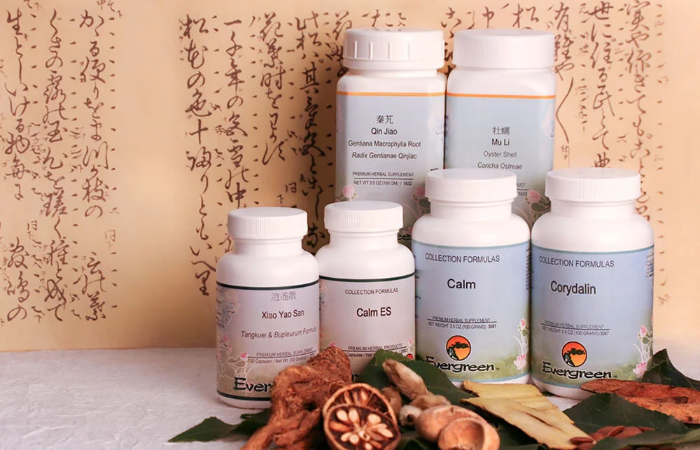 Six supplement bottles with labels from Evergreen herbal supplement brand, placed on a surface with dried herbs, roots, and sliced dried fruit in front of a background with handwritten Asian script.