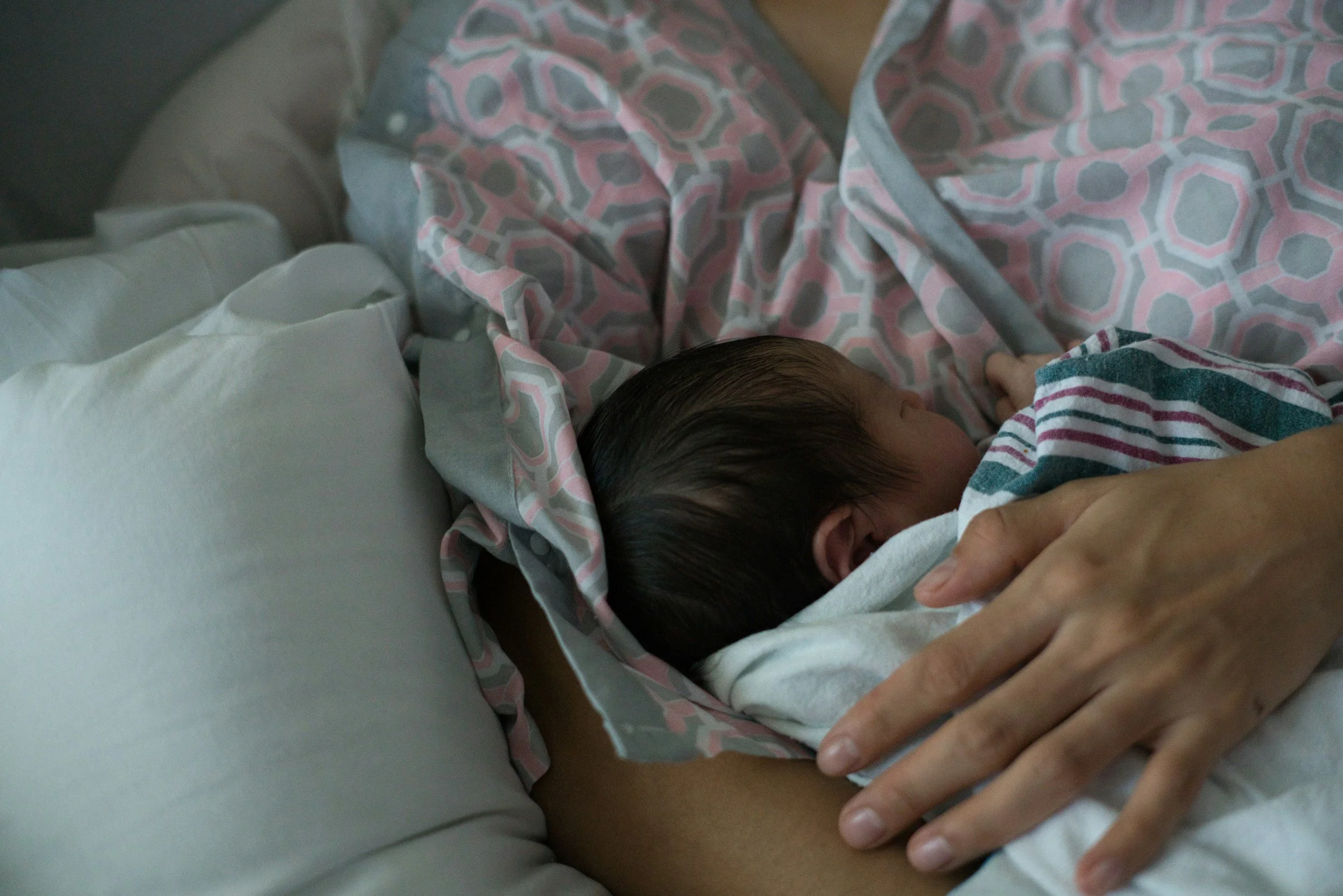 Close-up of a newborn baby sleeping on a person's chest, wrapped in a striped hospital gown, with an adult's hand gently resting on the baby's head.