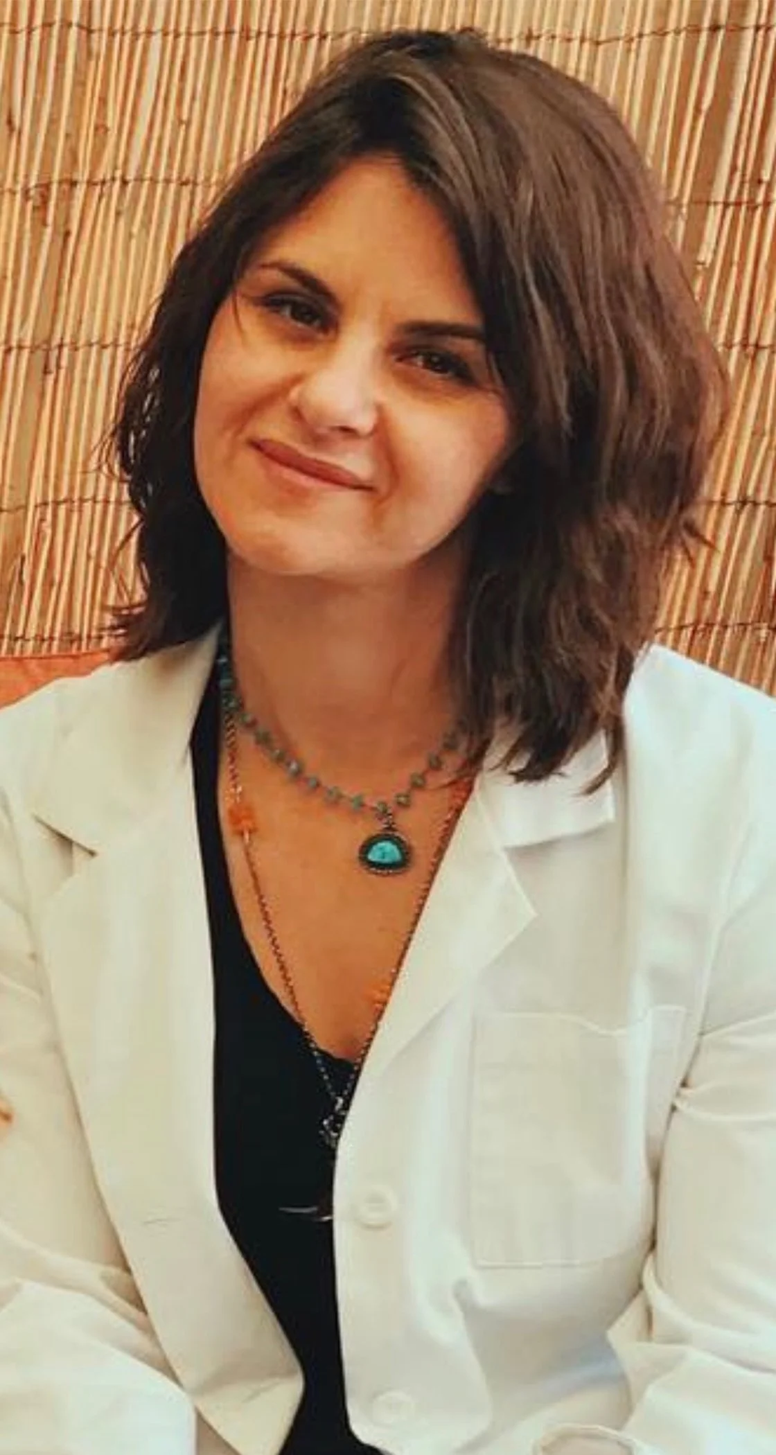 Woman with shoulder-length brown hair smiling, wearing a white blazer, layered necklaces including one with a turquoise pendant, in front of a bamboo background.