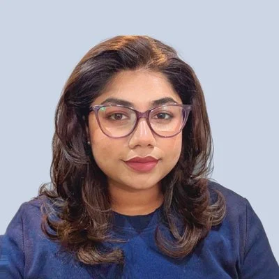 A woman with wavy brown hair, wearing glasses and a navy blue shirt, looking at the camera with a neutral expression against a light blue background.