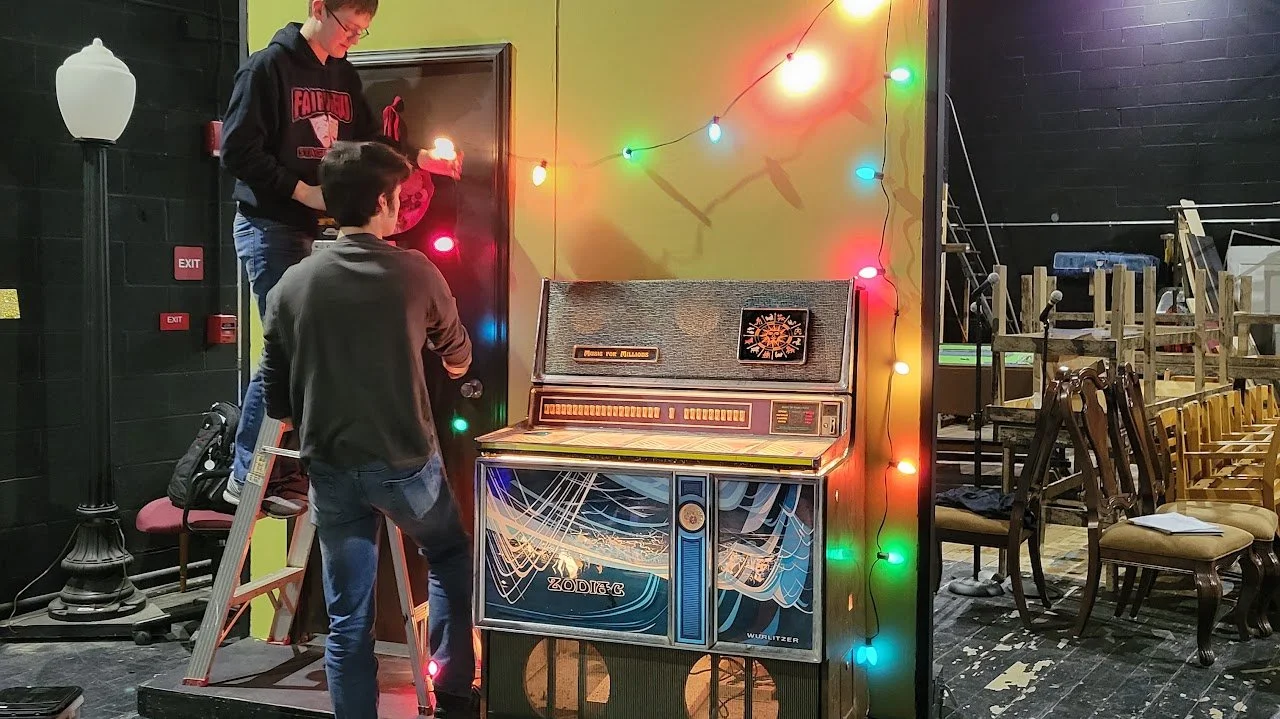 Two young men setting up or repairing a vintage arcade game machine decorated with multicolored string lights in a room with chairs and stage equipment.