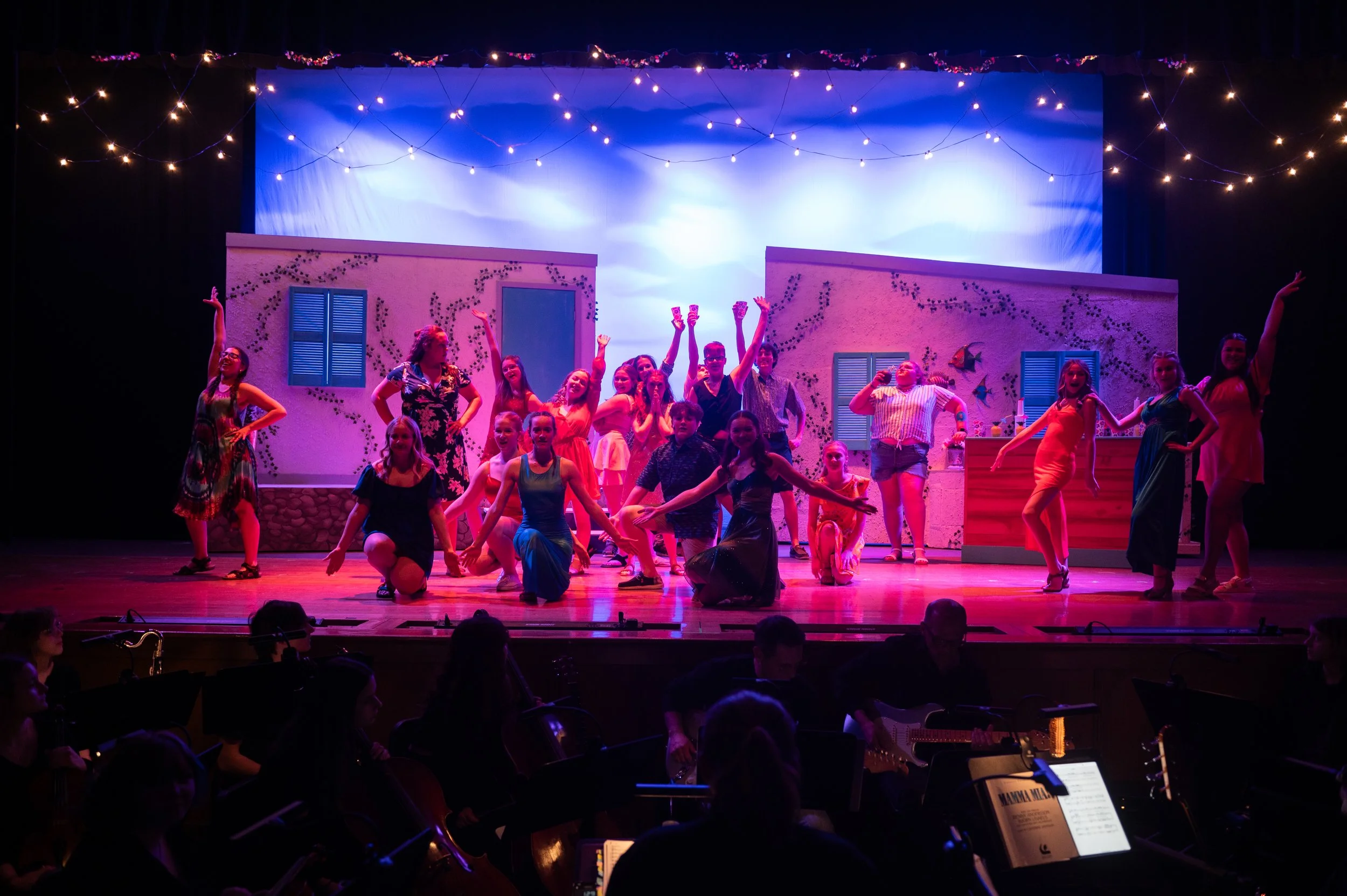 A group of performers on stage during a theater play, with some standing and others kneeling, smiling and striking various poses, in front of a windowed building backdrop, under string lights.