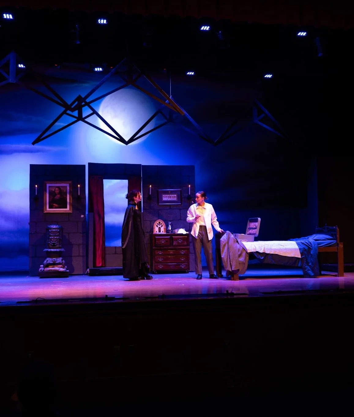Stage scene from a theatrical play featuring two actors in a bedroom setting with a bed, a dresser, and portrait paintings, illuminated by blue lighting.