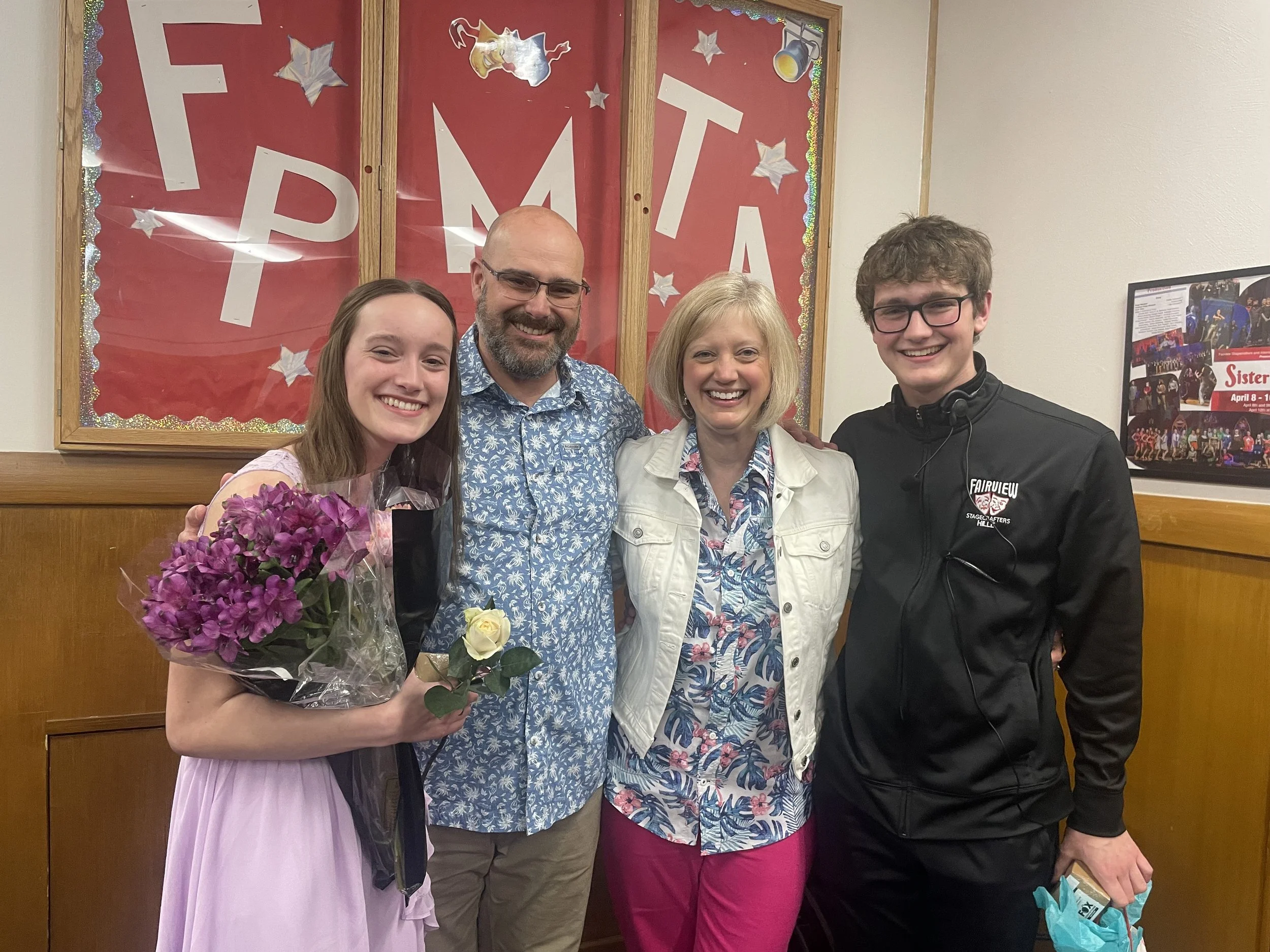 Four people standing together in front of a red bulletin board with the letters 'PROM' on it. The woman on the left is holding a bouquet of purple flowers and a white rose. They are all smiling.