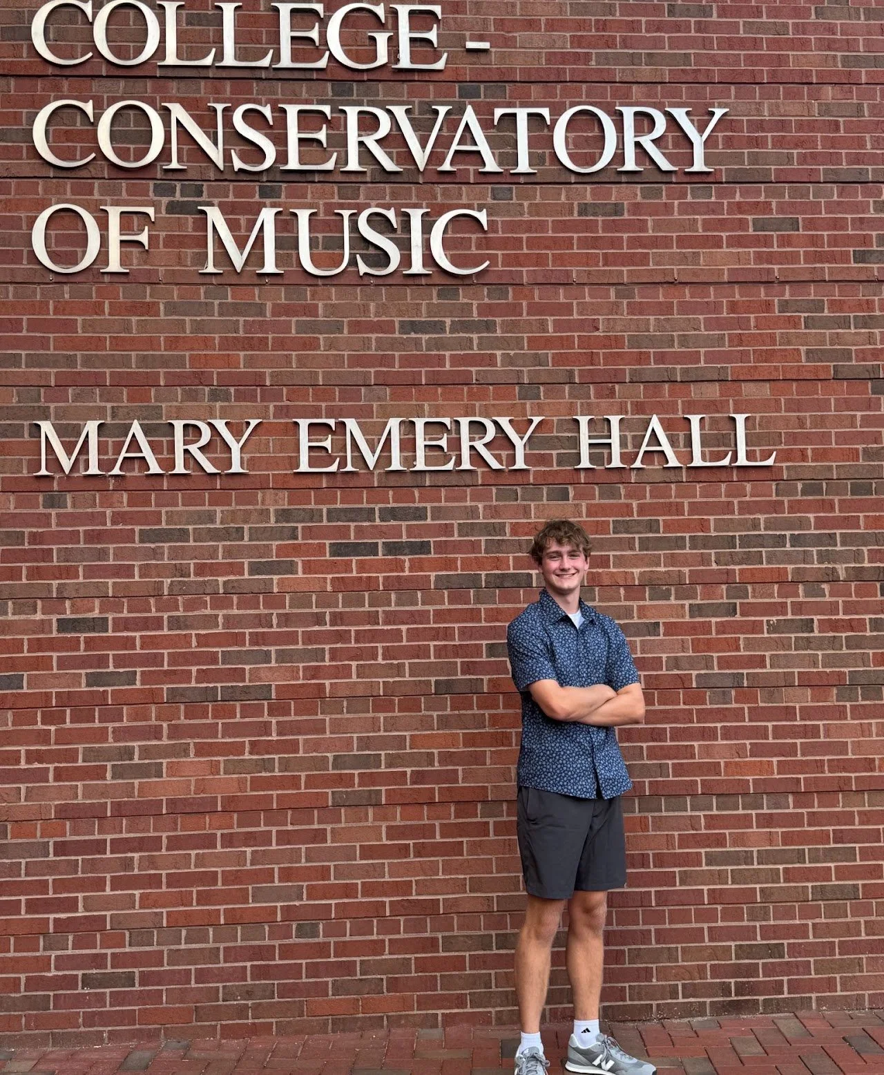 A young man standing in front of a brick wall with large white letters that read "College Conservatory of Music Mary Emery Hall." The young man is smiling with arms crossed, wearing a blue patterned short-sleeve shirt, black shorts, white socks, and 