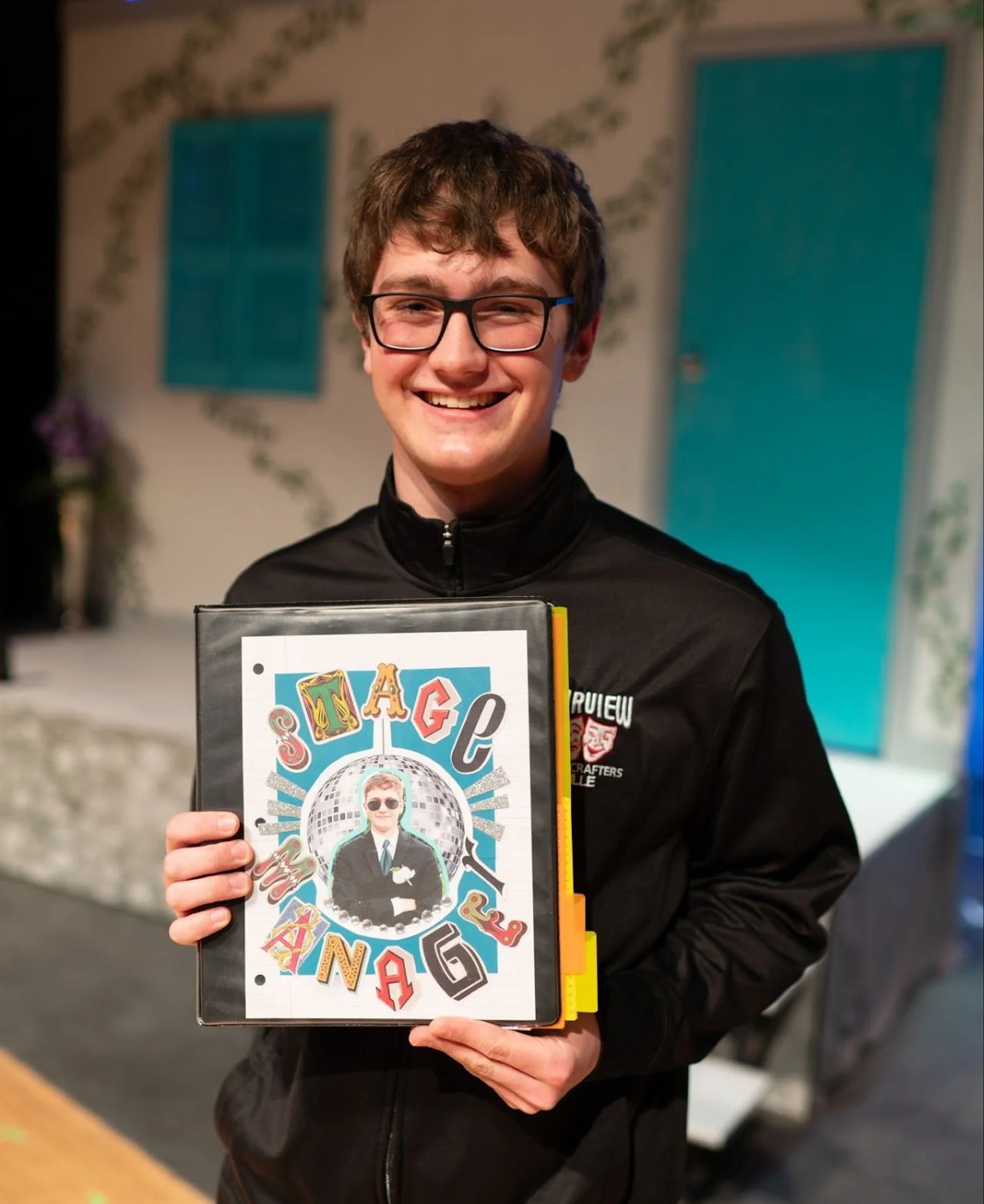A young man with glasses smiling and holding a book titled "Stag & Kout" with a vintage photo of a man in a tuxedo and sunglasses on the cover.