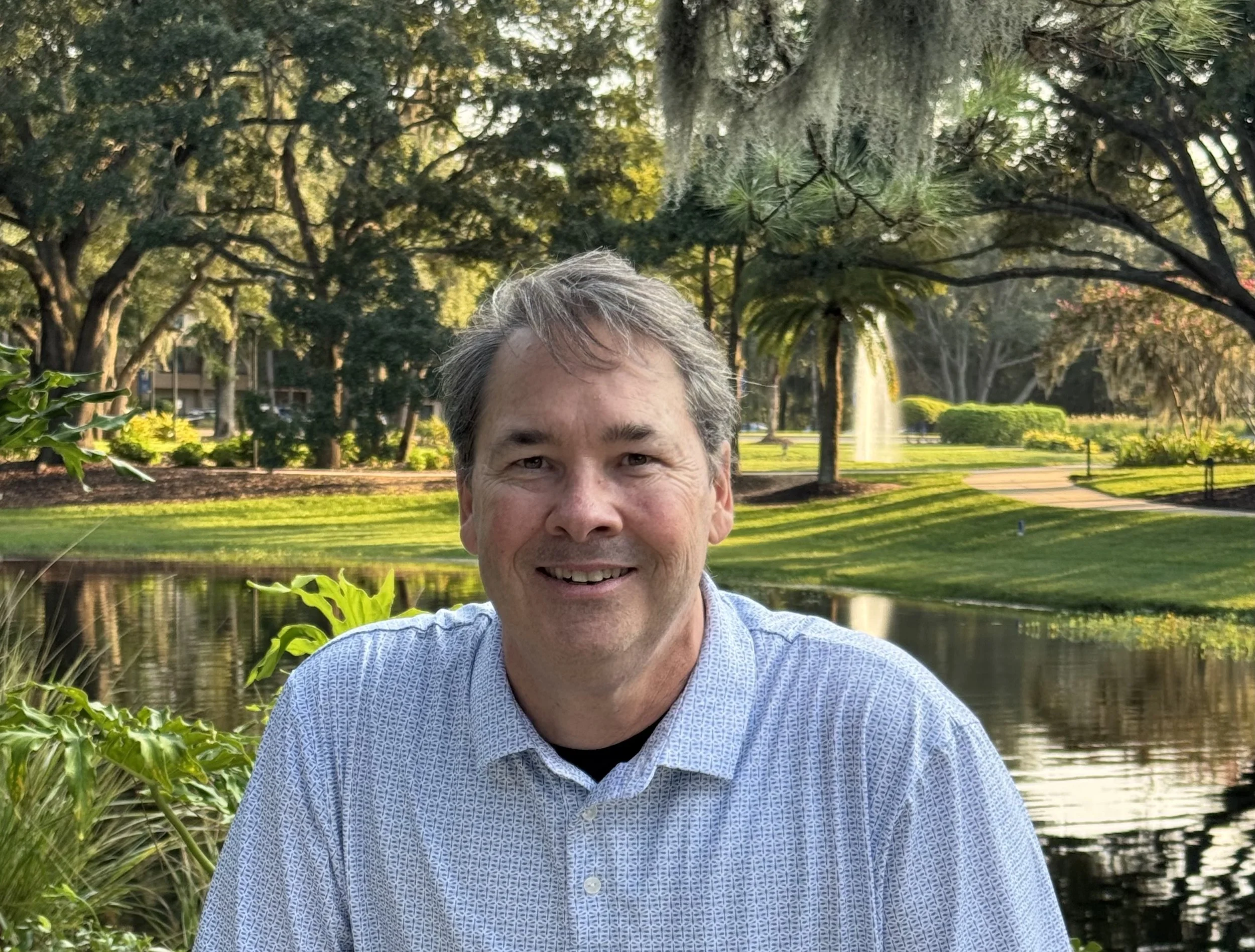 A middle-aged man with gray hair smiling in front of a park with trees, a fountain, and a pond.