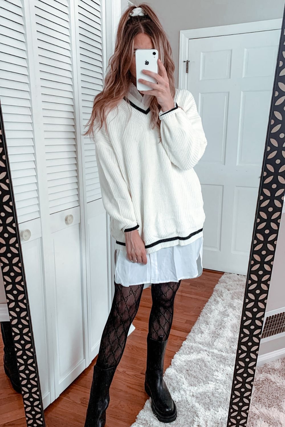 woman taking a mirror selfie wearing a white and black oversized sweater over a white shirt dress paired with black patterned tights and black mid thigh rubber boots