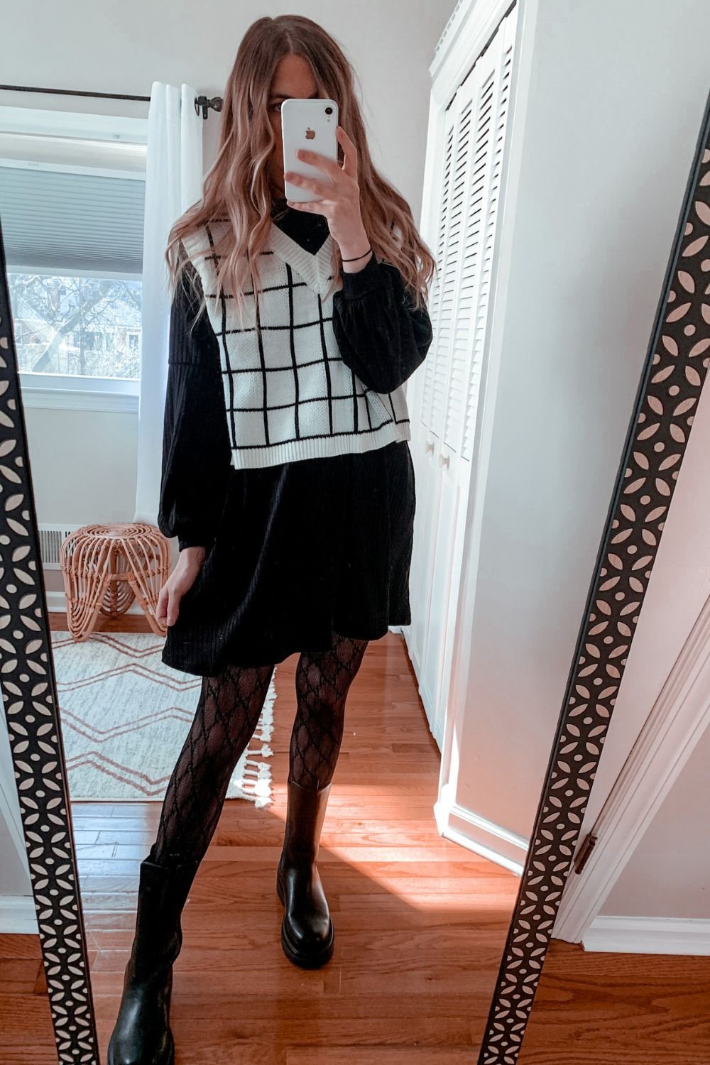 woman taking a mirror selfie wearing a white and black plaid cropped sweater vest over a black long sleeve flowy dress paired with black patterned tights and black mid thigh rubber boots