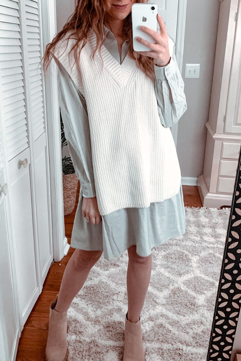 woman taking a mirror selfie wearing a light beige sweater vest over a light green shirt dress paired with beige ankle boots