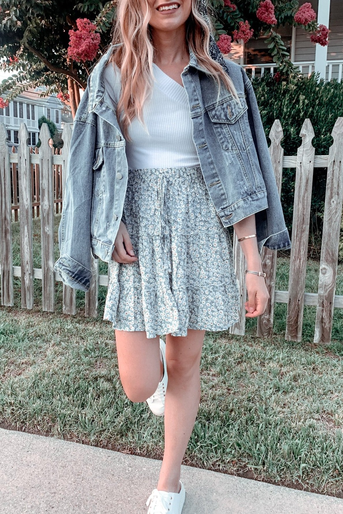 woman on the sidewalk wearing a denim jacket over a white top and green floral mini skirt with white sneakers