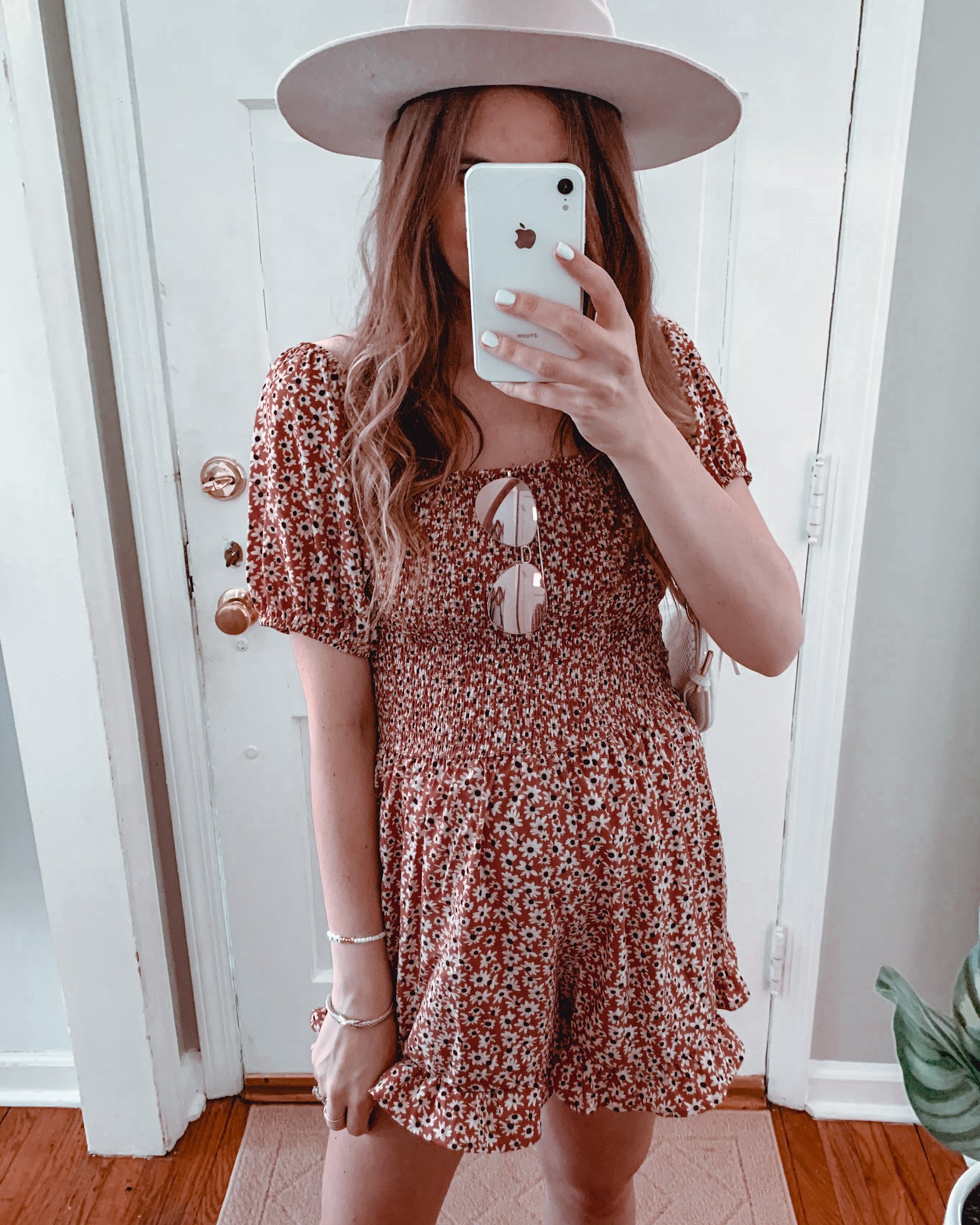 woman taking mirror selfie wearing a beige wide brim fedora hat, red floral romper and pink tinted sunglasses