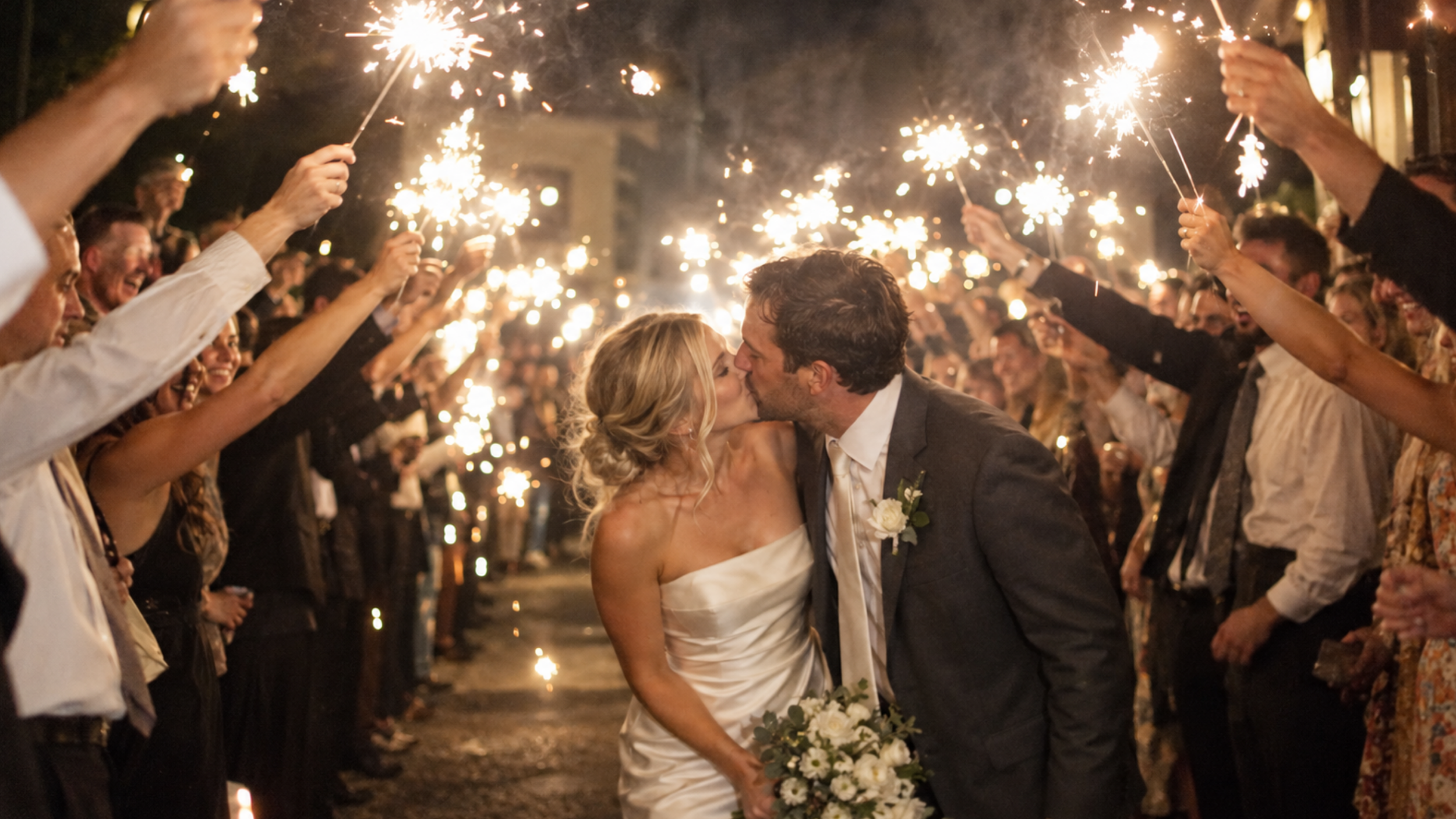 Newlyweds kissing beneath raised sparklers, with the bride wearing a strapless satin gown.