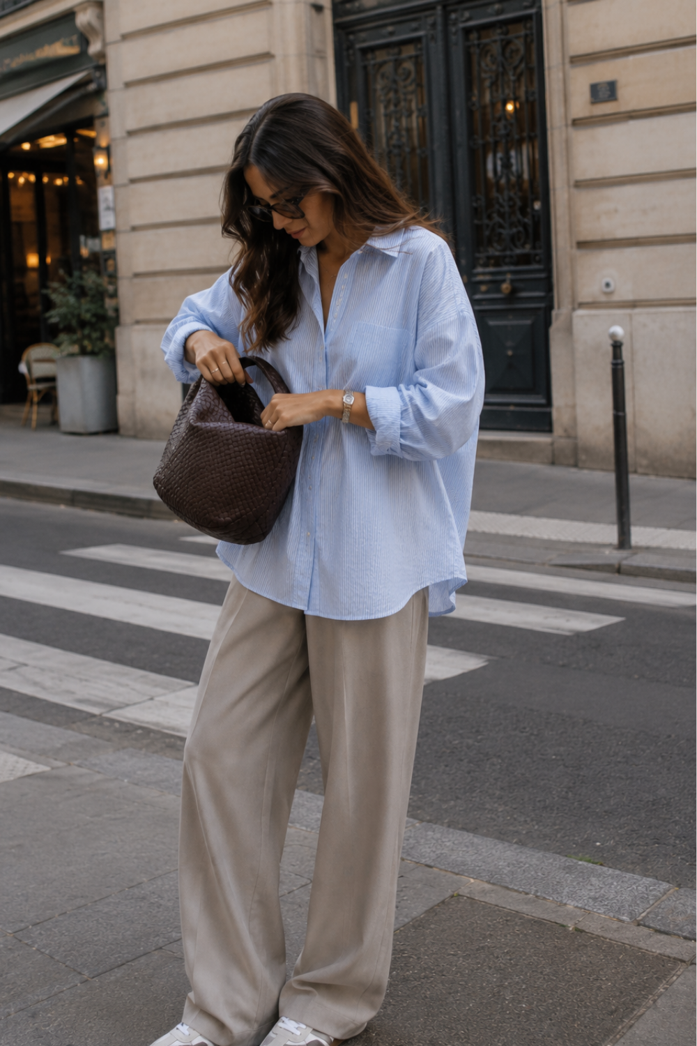 trendy woman wearing a blue button down and trousers looking into her purse on the street