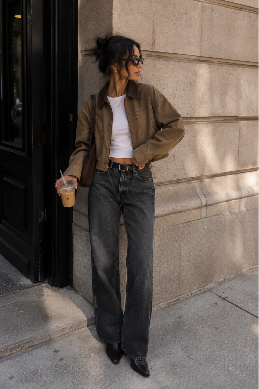 trendy woman wearing a brown cropped jacket and jeans leaning against a building