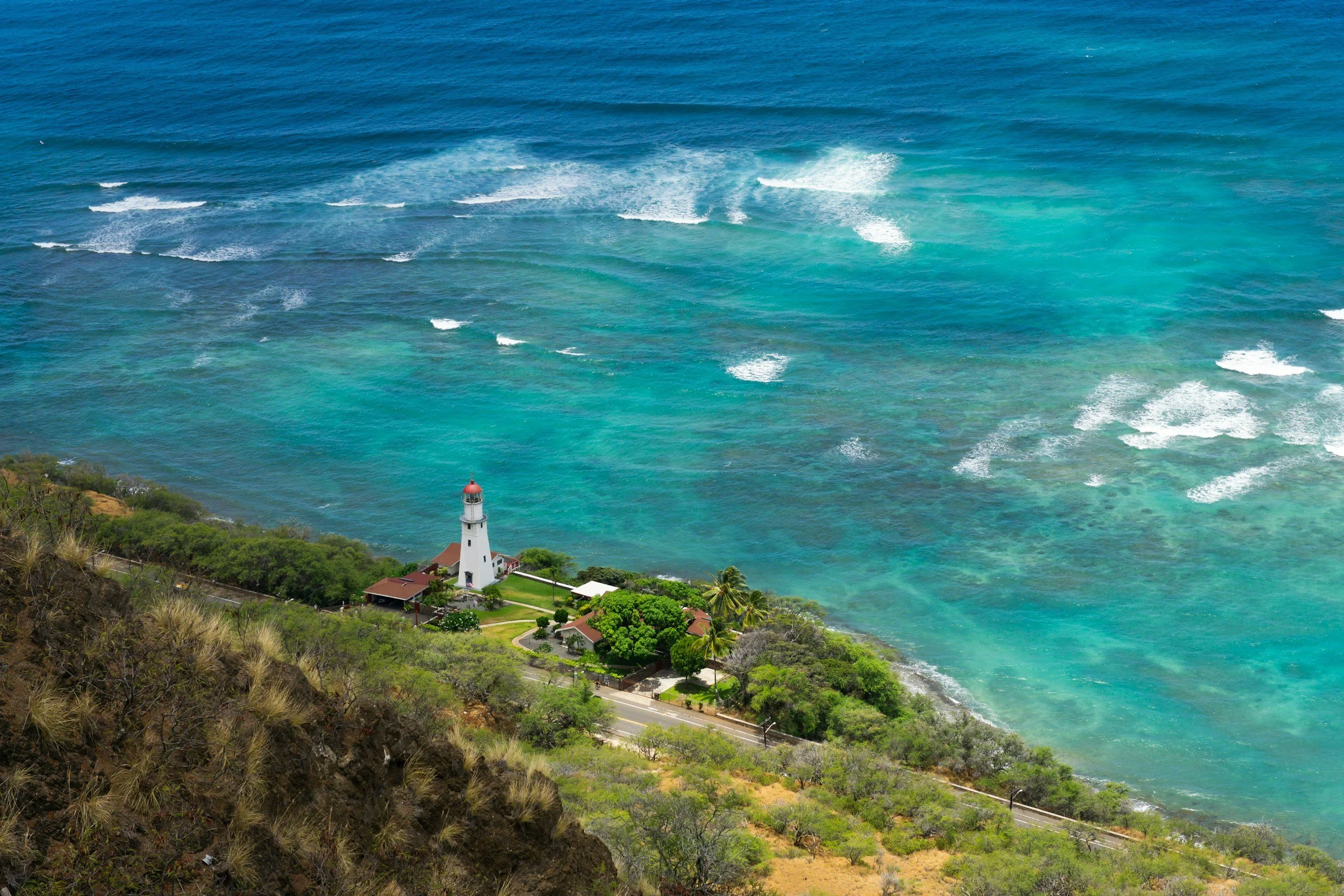 A lighthouse on a green hillside overlooking the ocean, with waves and clear blue water.