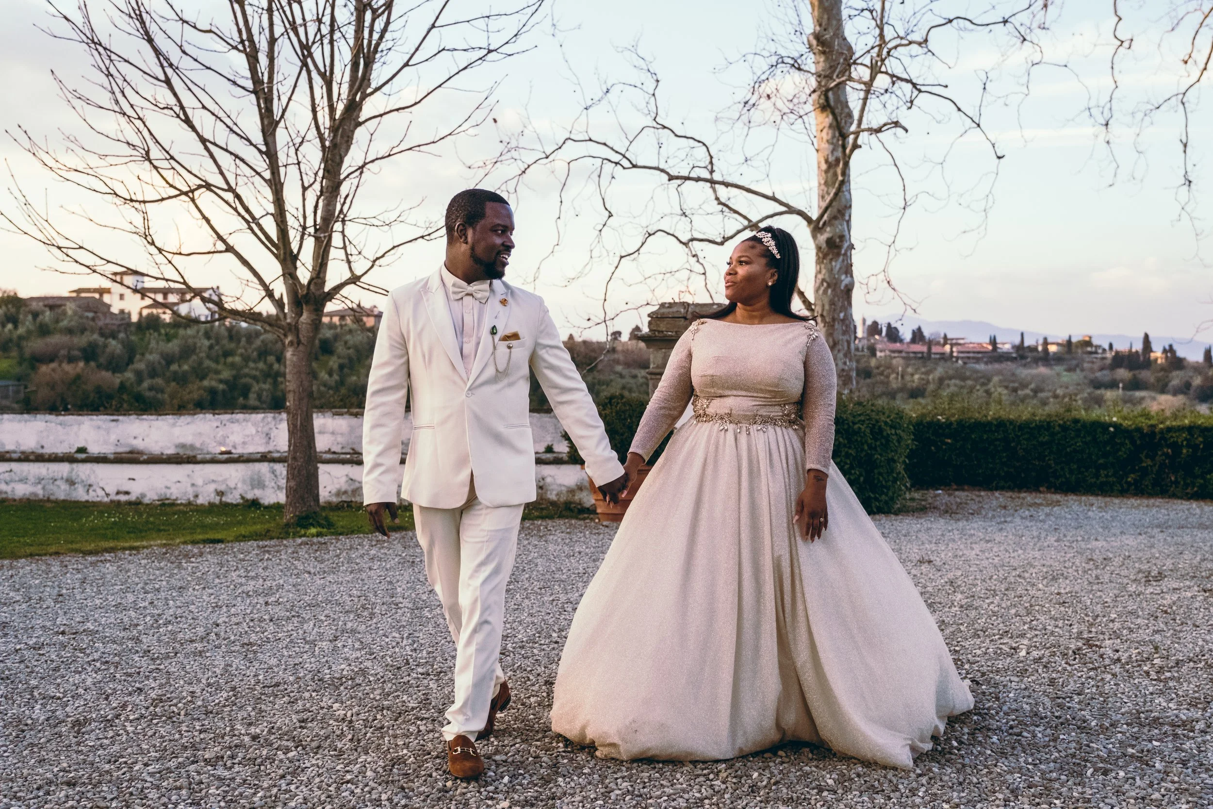 A couple in wedding attire holding hands, walking outdoors on gravel, with leafless trees and distant landscape background.