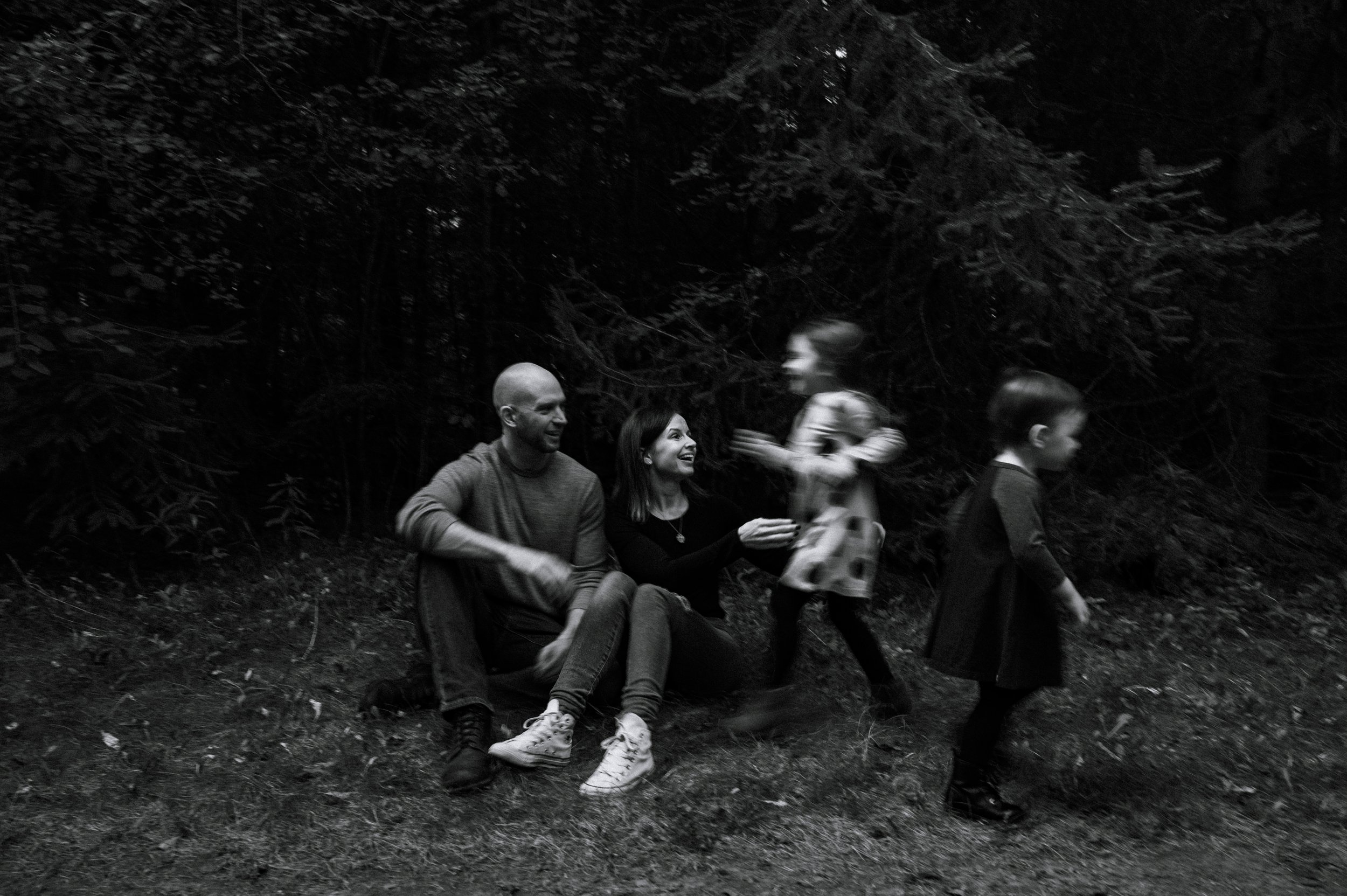 Parents sit on a grassy patch of a trail, with trees behind them, as their two daughters fun around, so fast that they're blurry, during a family photo session in Newmarket, Ontario.