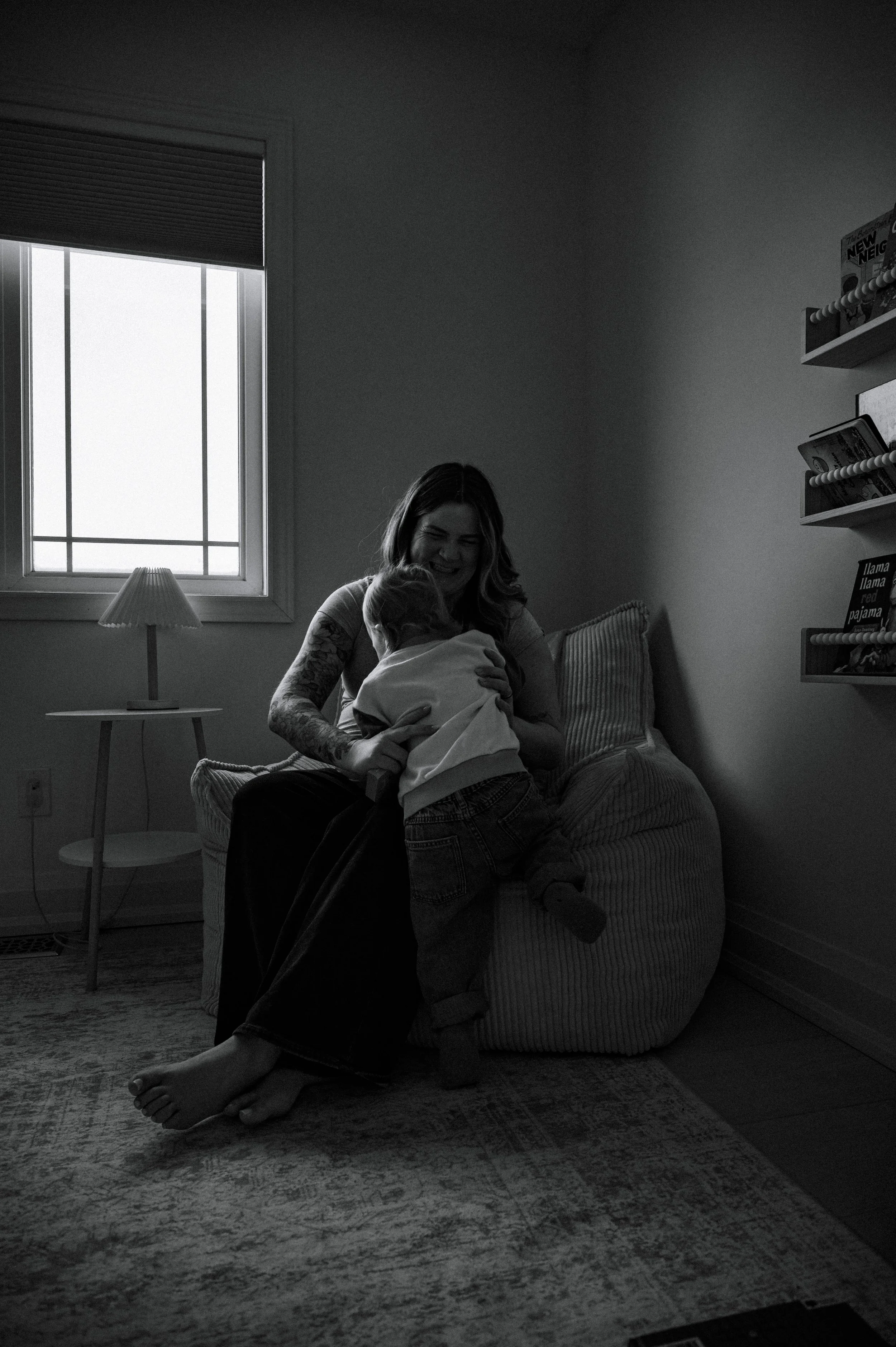 A mom sitting in a comfy chair, leaning over to pick up her fast-moving toddler who has brought her a book to read, during their in-home family photo session in Georgina, Ontario.