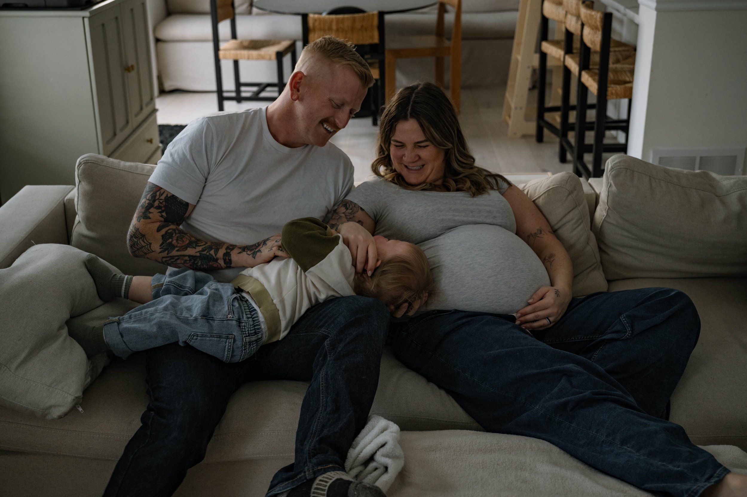 A father and mother laughing together with their toddler, all cuddled up, during a lifestyle in-home maternity session.
