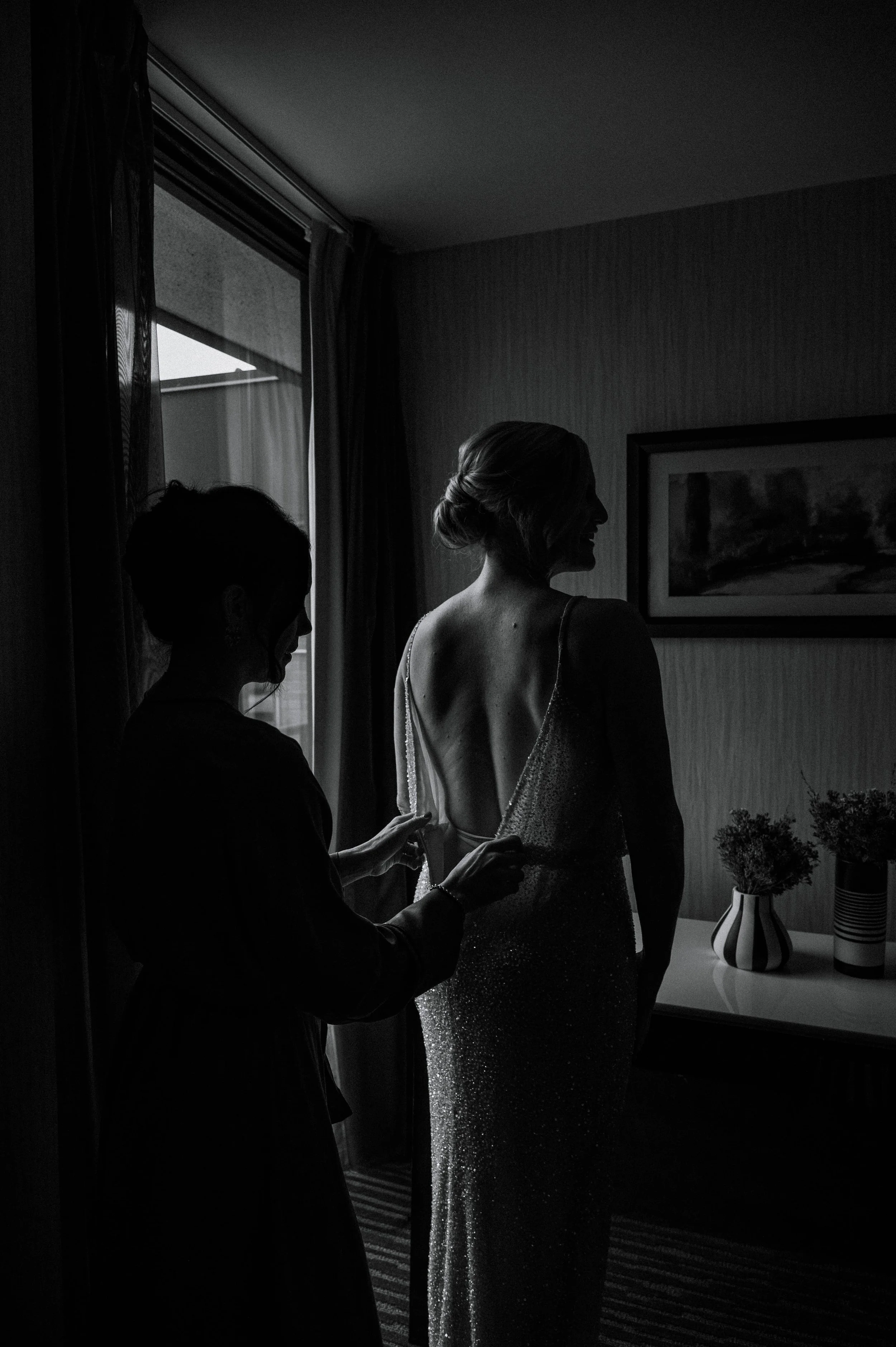 Documentary wedding photography in Hamilton, Ontario: a dramatic, almost silhouette, portrait of a bridesmaid doing up the bride's sequin wedding dress.