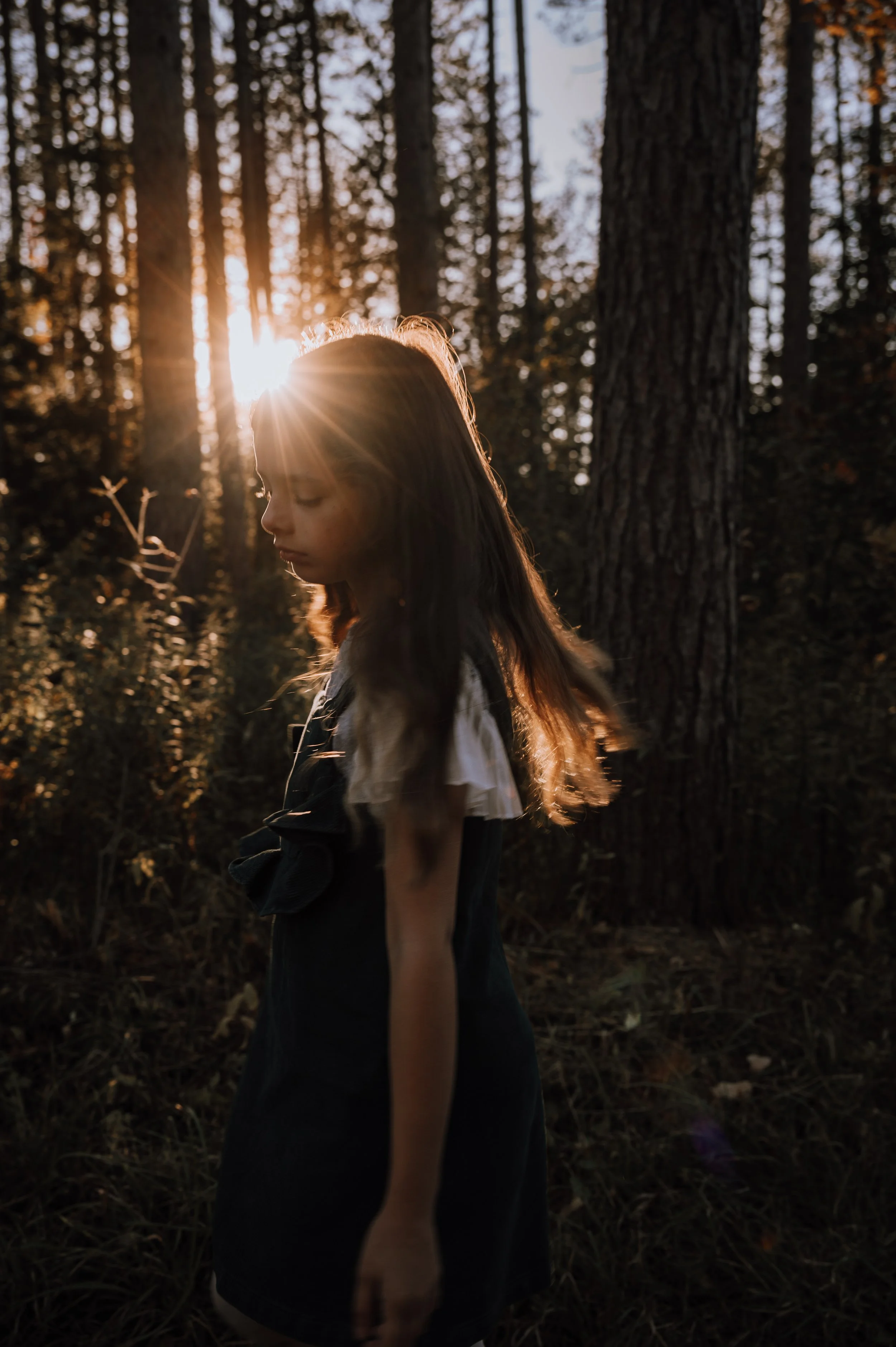 A young girl spinning in circles in a forest as the sun peeks through the trees in a perfect sun flare over her head, during a family session in Newmarket.