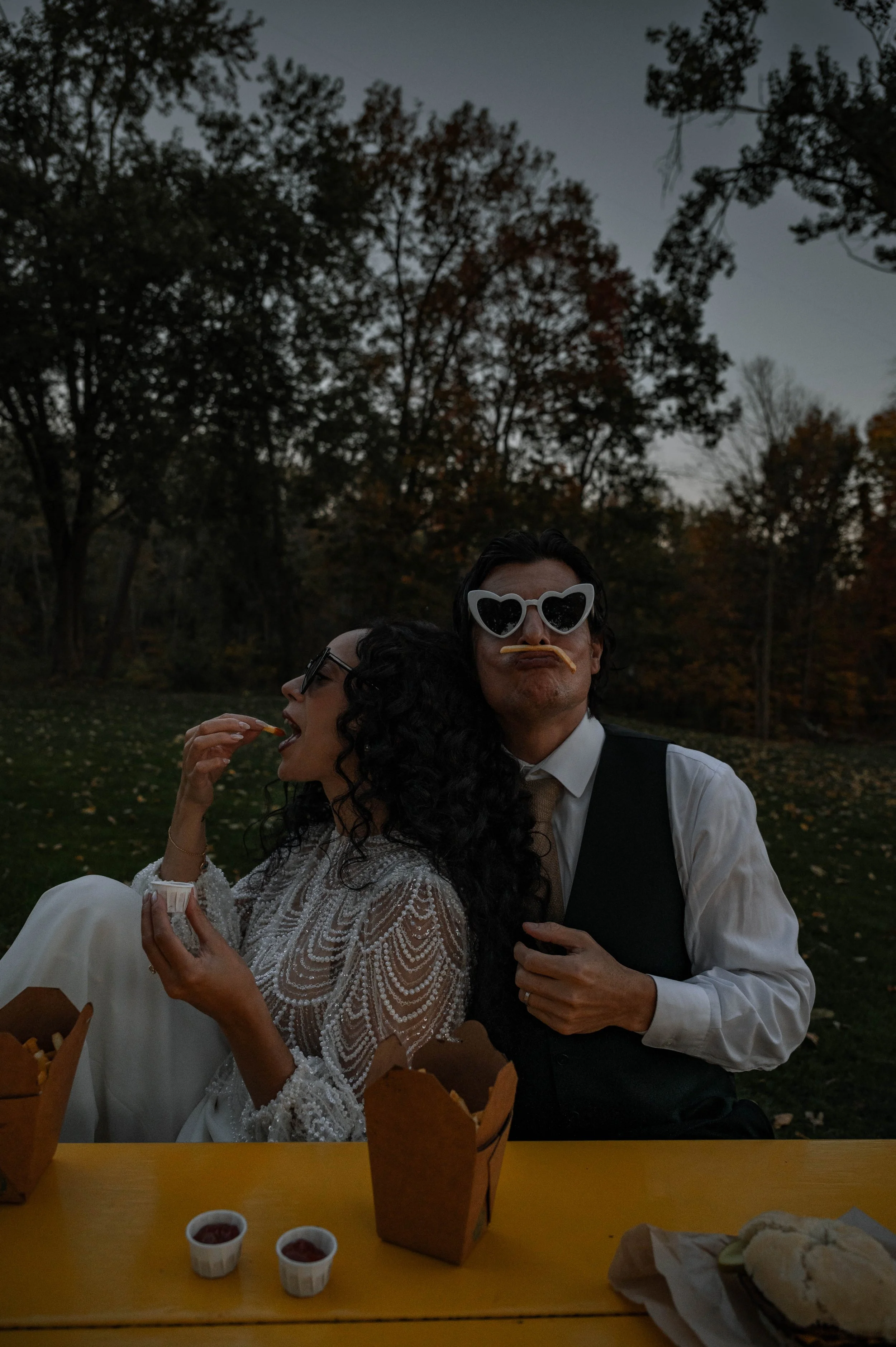 A bride and groom eating french fries together, wearing heart sunglasses. The groom has a fry as a moustache. During a wedding elopement in Orillia, Ontario.
