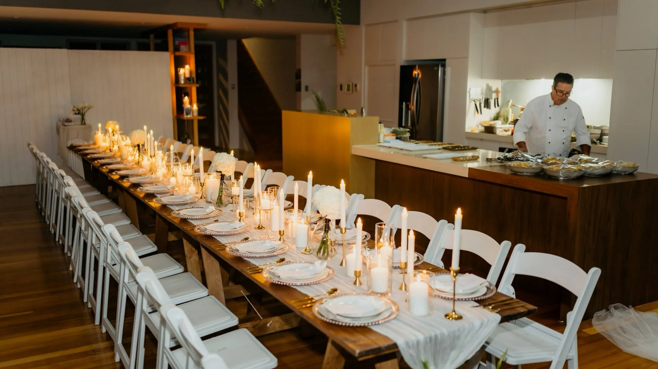 Candlelit long table set for a private dinner at home, with a Michels chef in the kitchen