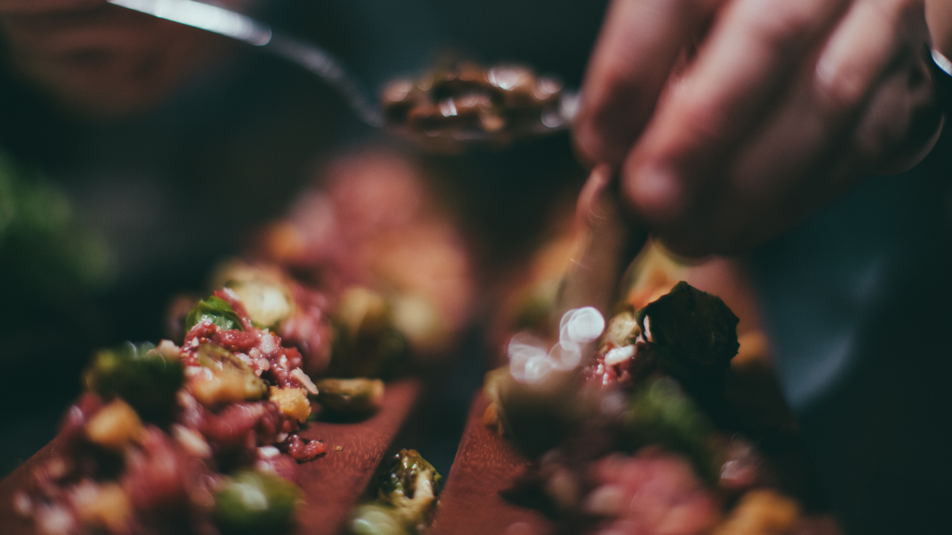 Close-up of a person’s hand scooping food from a serving dish, with food on a plate and a blurred background.