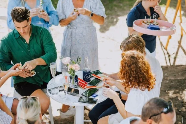 People gathered around a white table outdoors celebrating with food and drinks, some taking photos or using their phones.