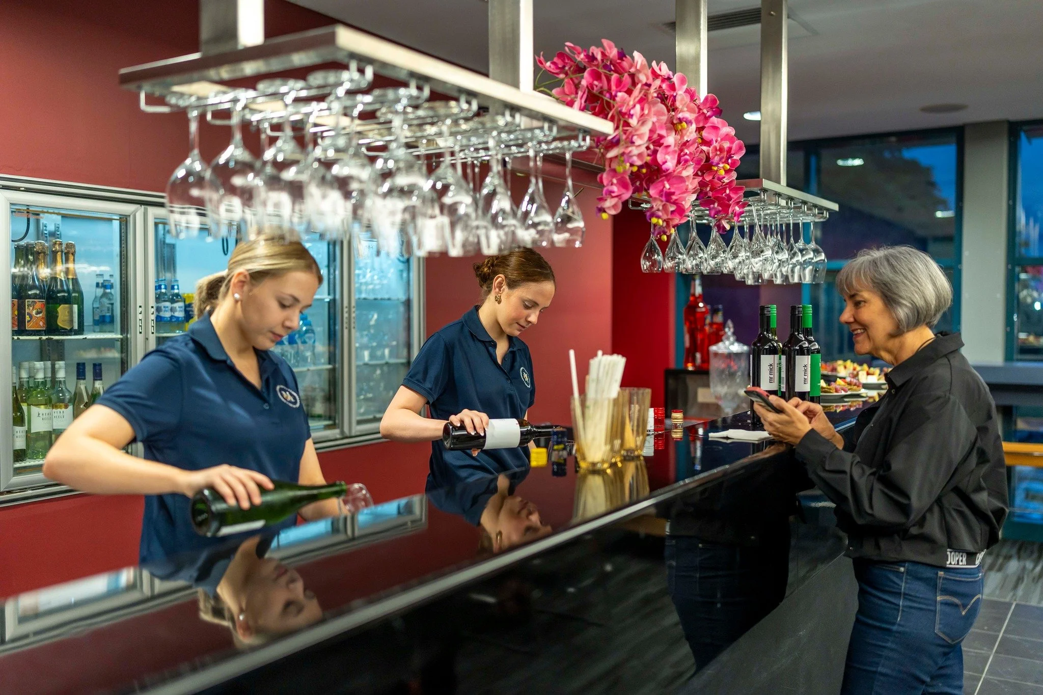 A woman at a bar ordering a drink, with two female bartenders preparing beverages behind the counter. The bar has glassware hanging above and bottles of alcohol behind glass refrigerators.