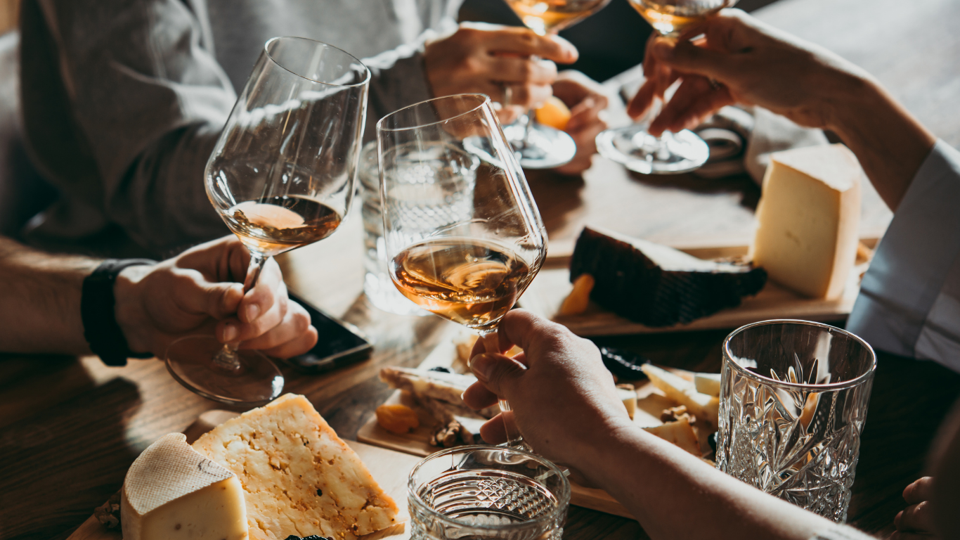 People toasting with glasses of rosé wine at a wooden table with cheese, crackers, and charcuterie.