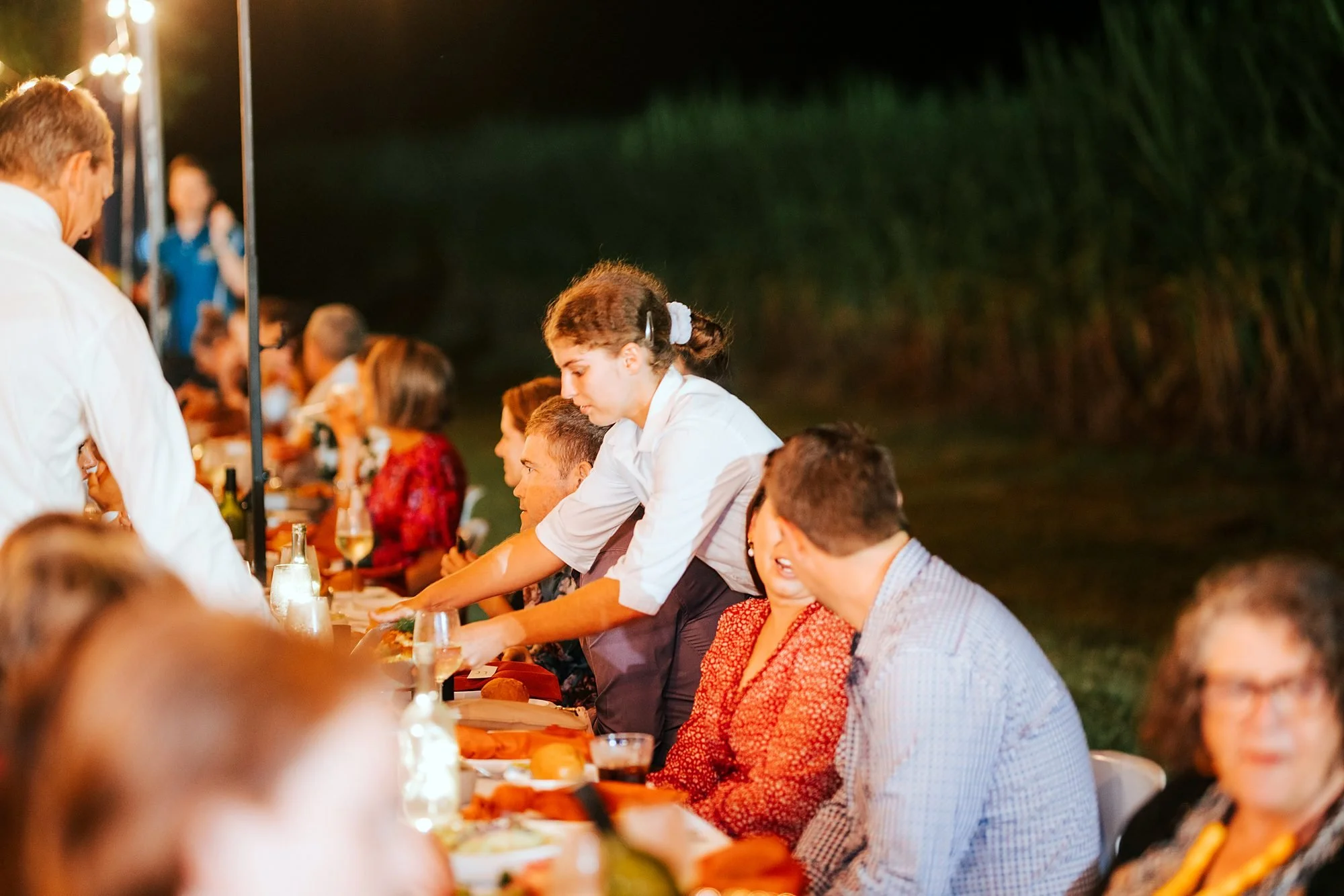 Michels waitstaff serving guests at a long-table dinner under festoon lights
