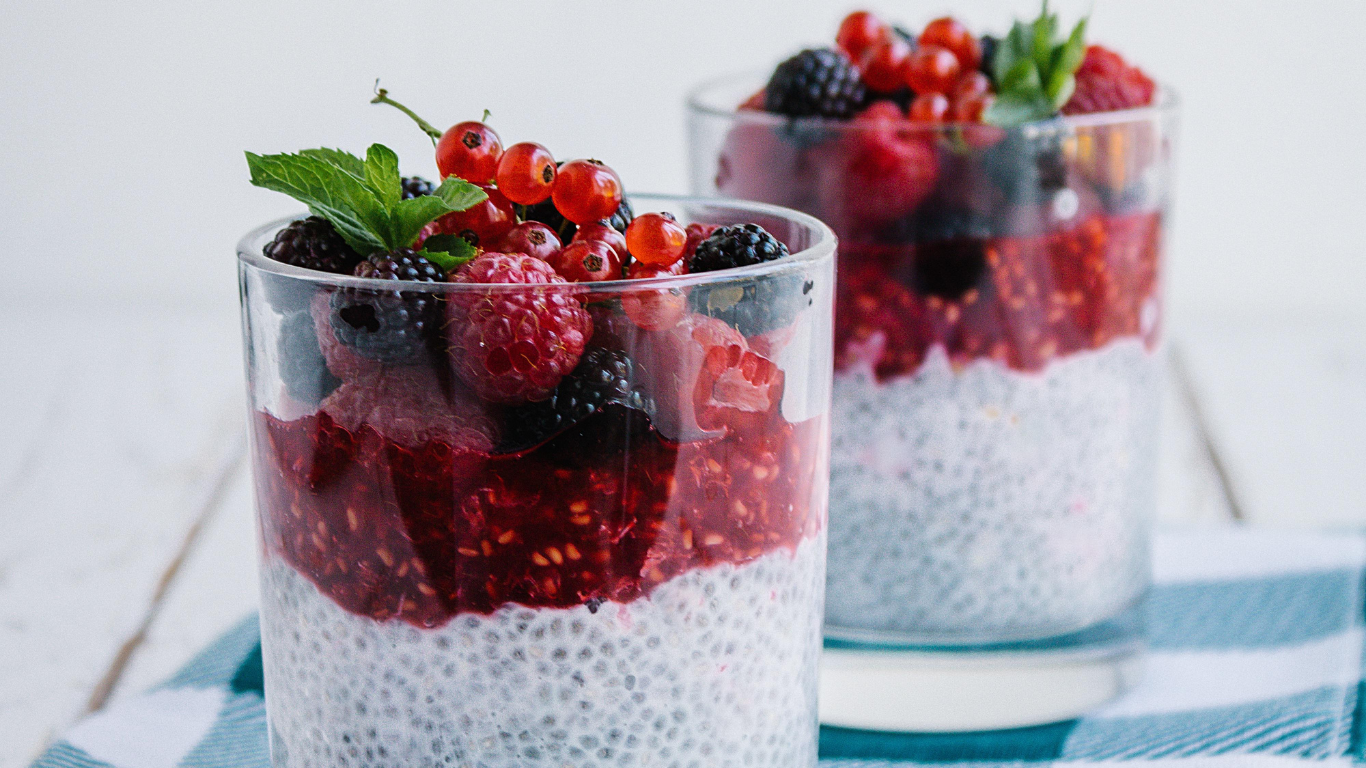 Two glasses filled with chia seed pudding topped with mixed berries, red currants, and fresh mint leaves, placed on a blue and white striped cloth.