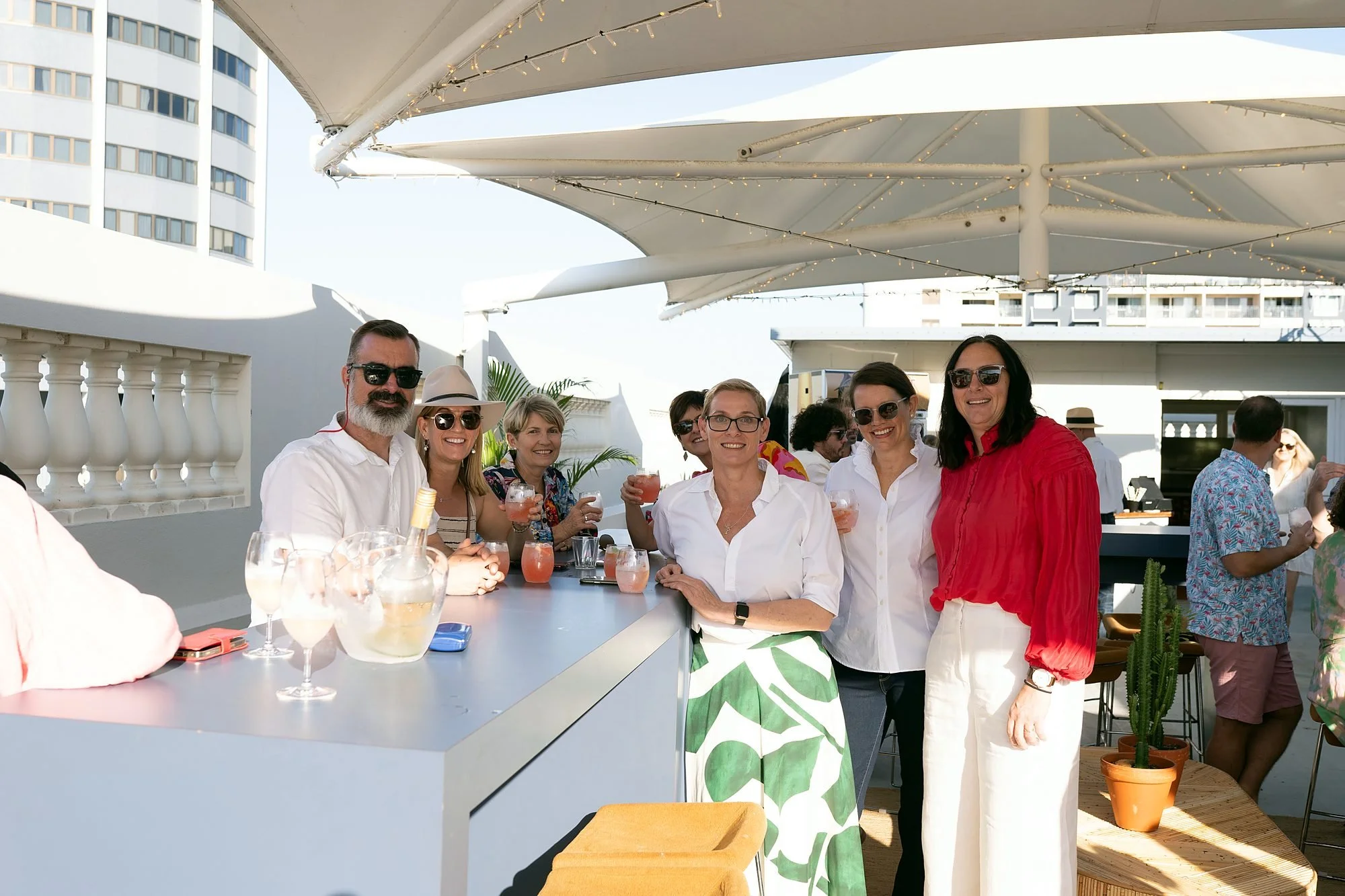 Smiling guests with drinks at a rooftop bar under white shade sails