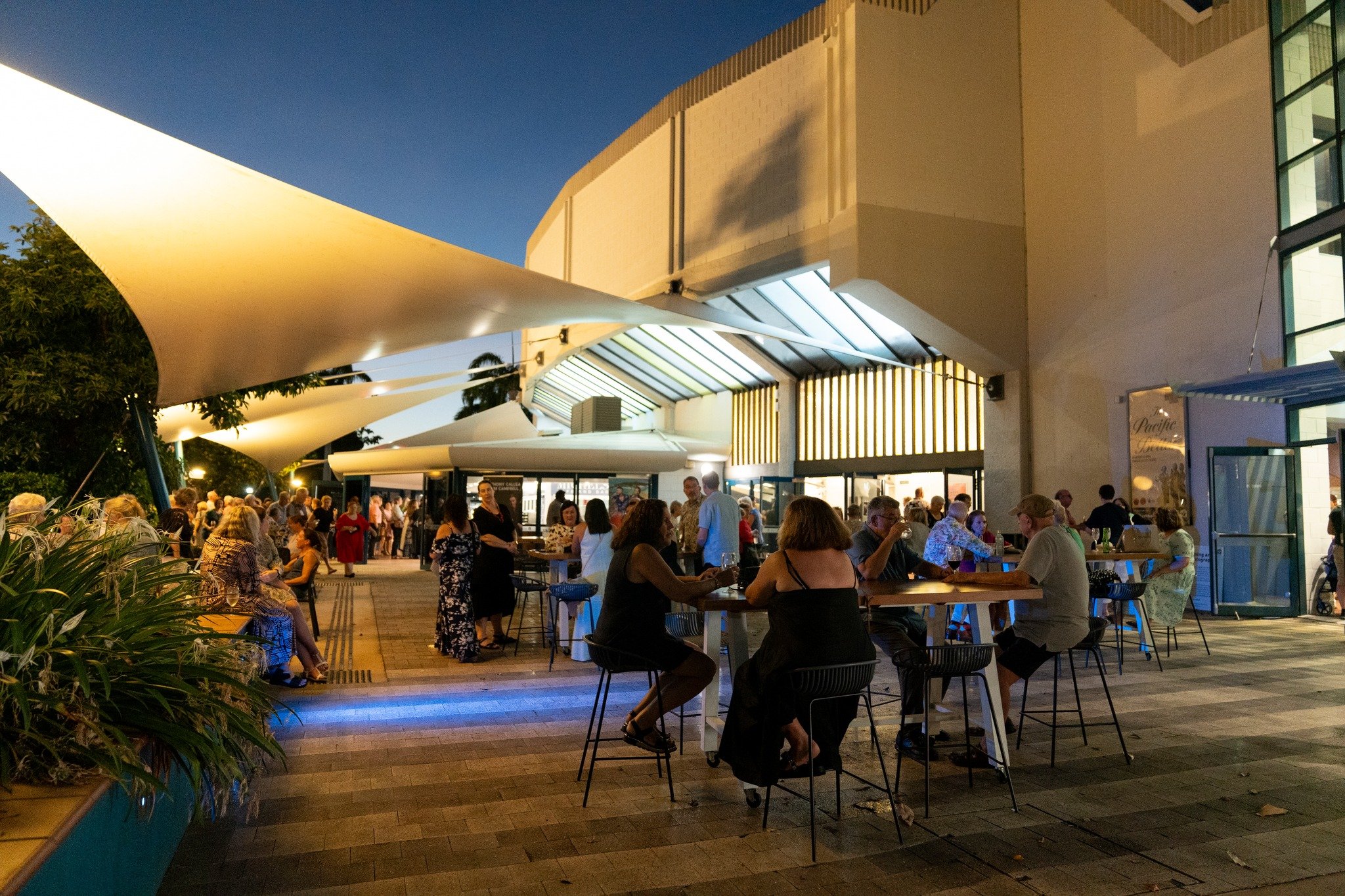 People dining and socializing at outdoor tables under large shade structures at dusk, near a modern building with illuminated windows.