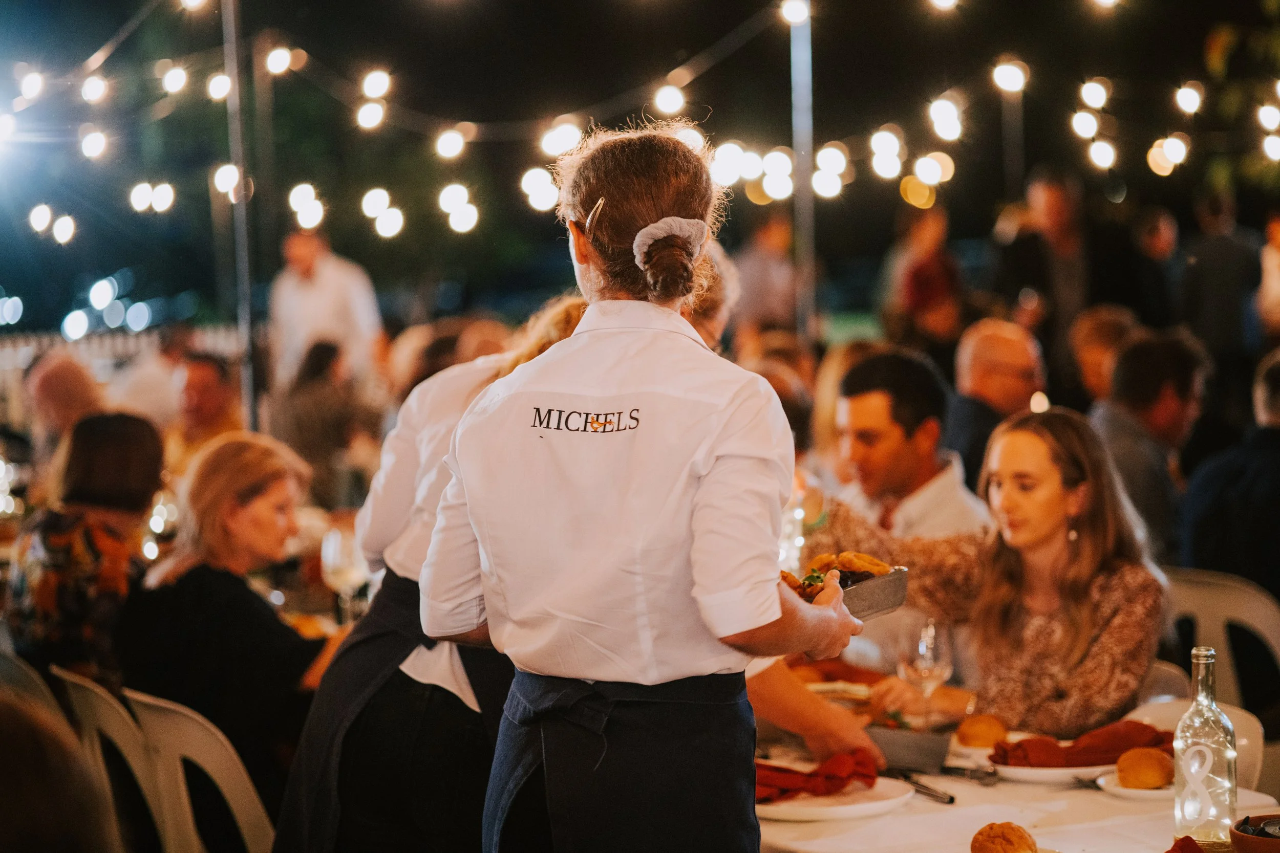 A woman in a white shirt with the logo 'MICHELS' on the back is serving food at an outdoor evening event with string lights overhead and people seated at long tables.
